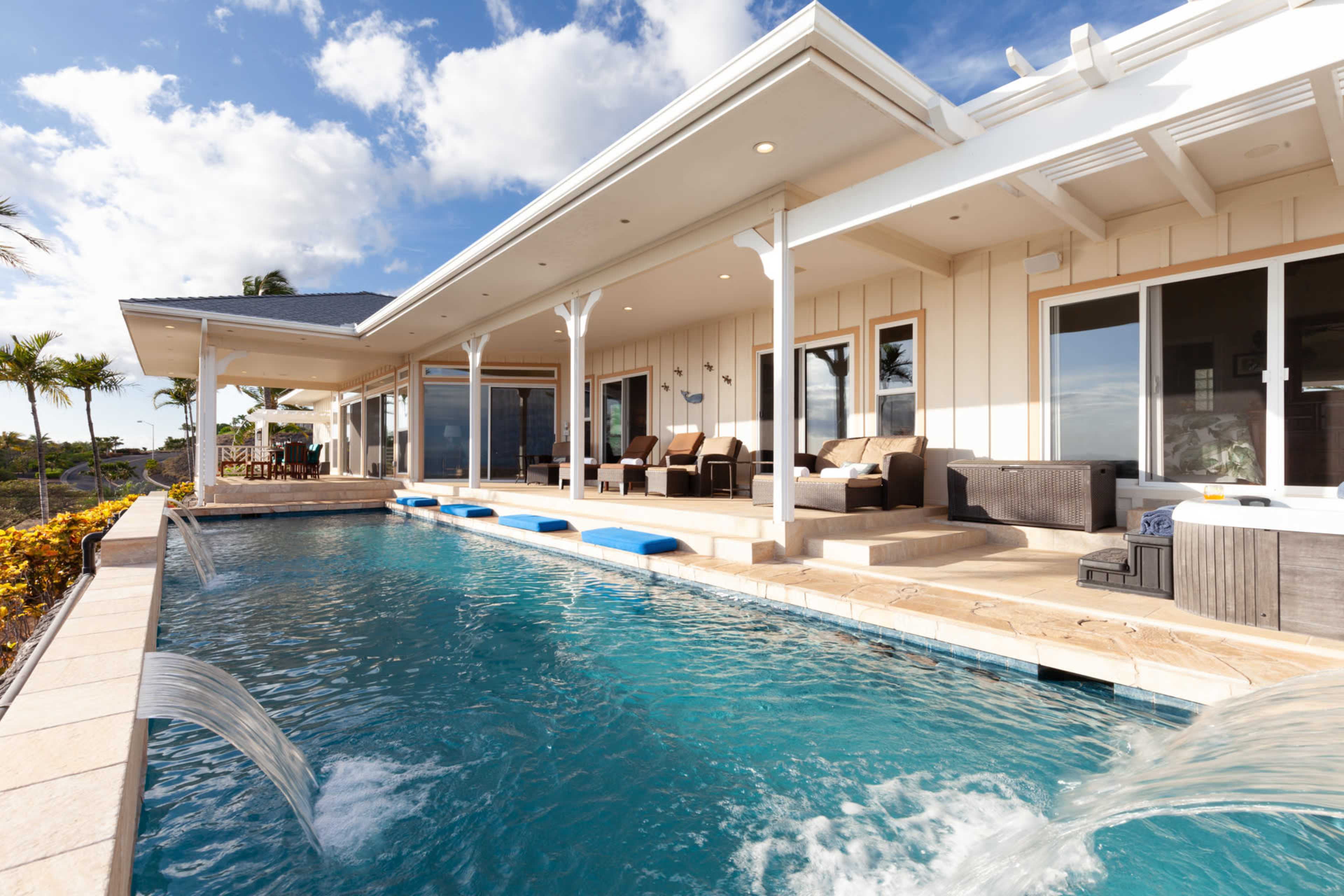 A modern villa features a swimming pool with water fountains, surrounded by outdoor seating and palm trees.