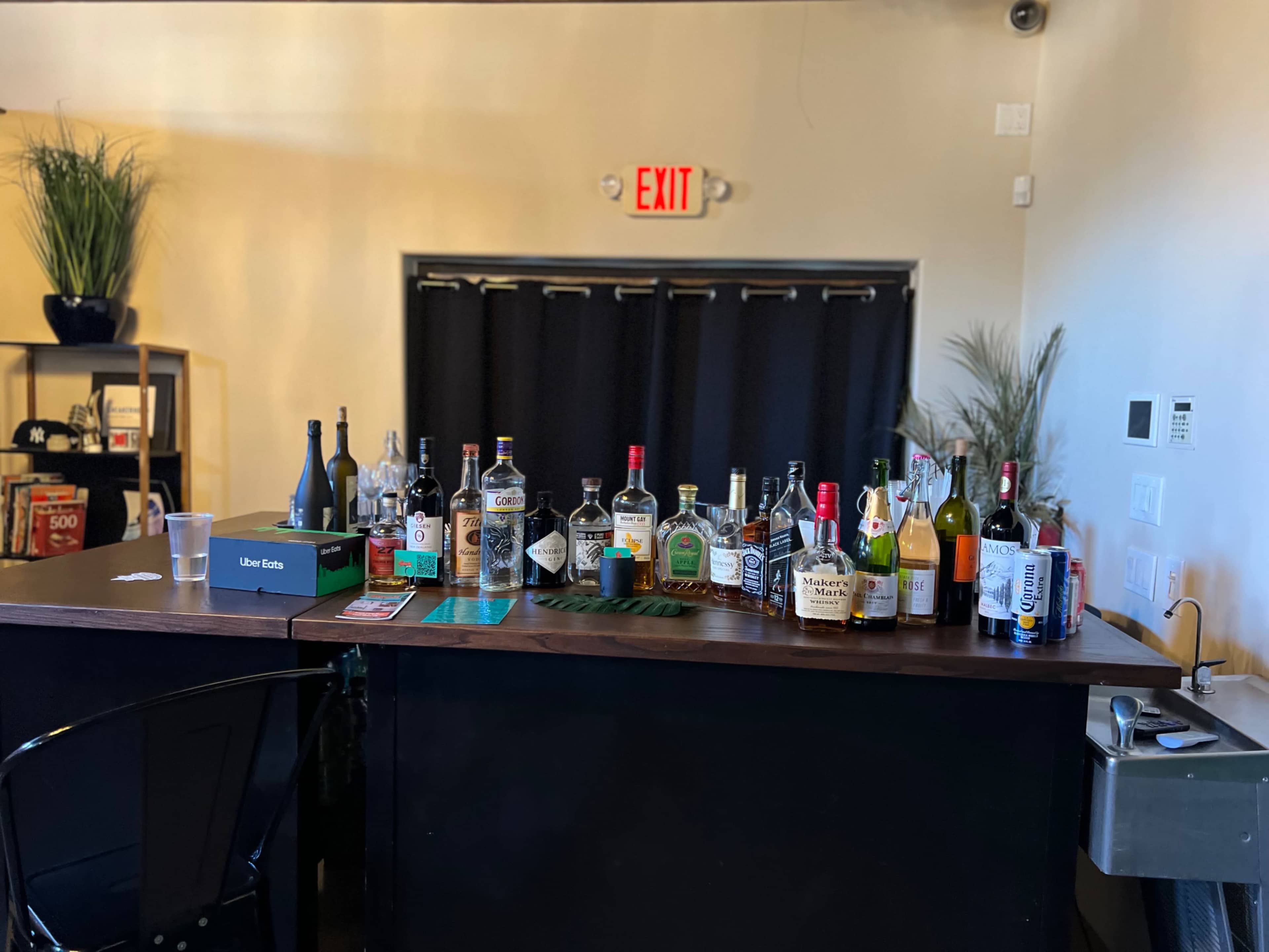 The image shows a bar setup with various bottles of liquor displayed on a wooden counter, behind which there is a black curtain and an exit sign.