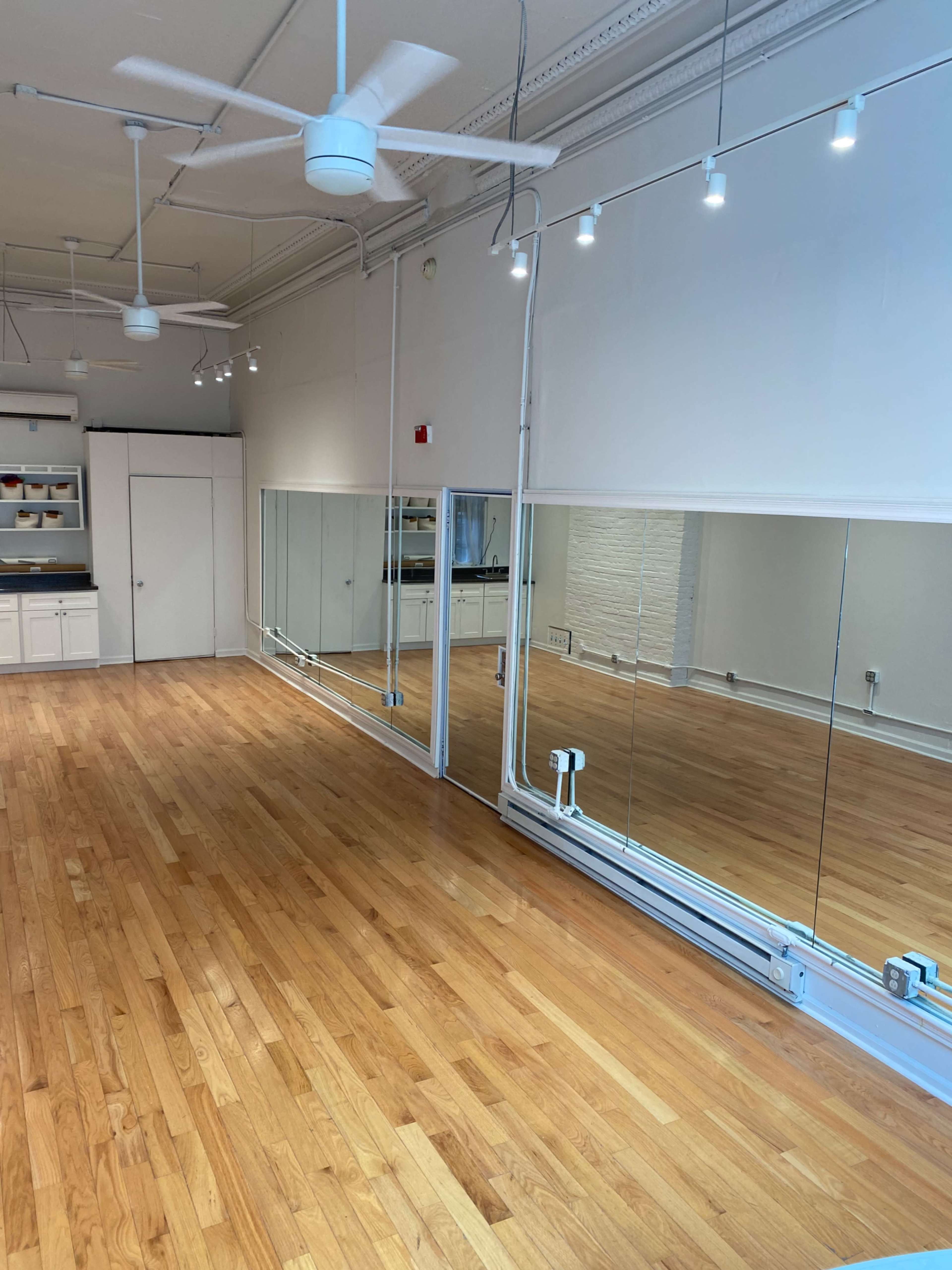 A spacious, well-lit room with wooden floors, large mirrors on one wall, and overhead lighting fixtures.