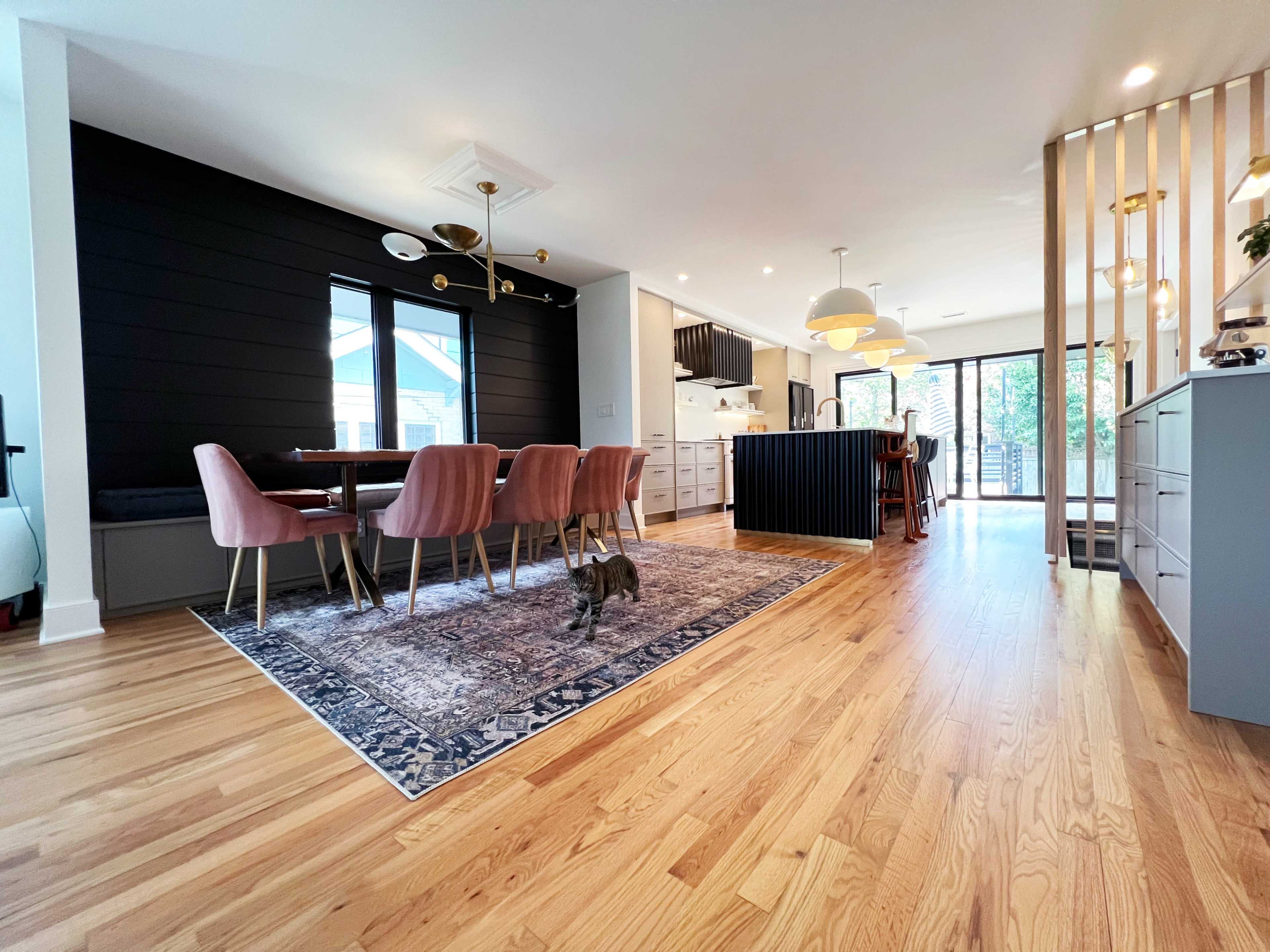 A modern open-concept kitchen and dining area featuring a large dining table with pink chairs, a dark island counter, and wooden flooring.