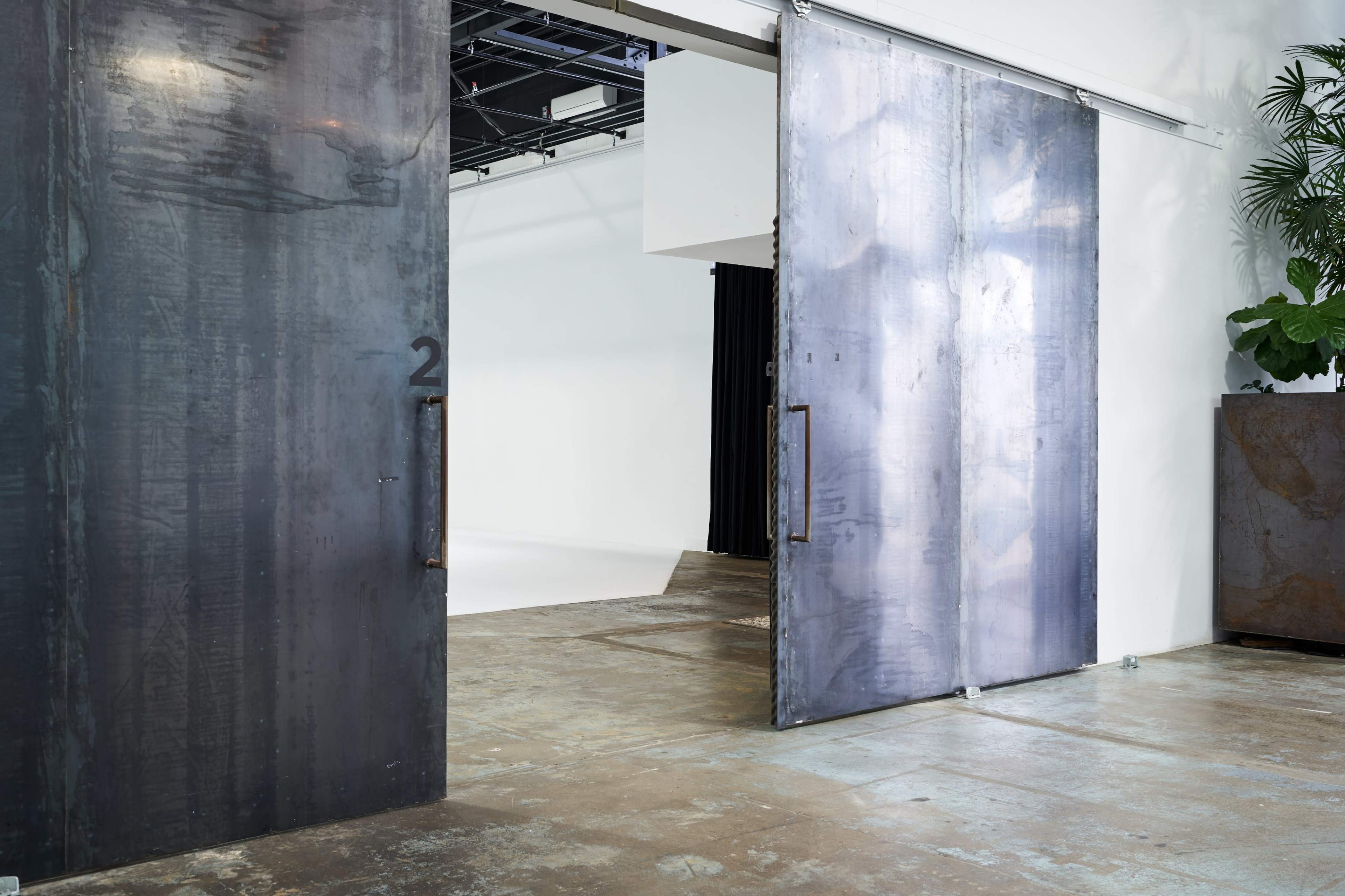 Large metal sliding doors slightly open, revealing a blank white studio space behind them, with a potted plant in the foreground.