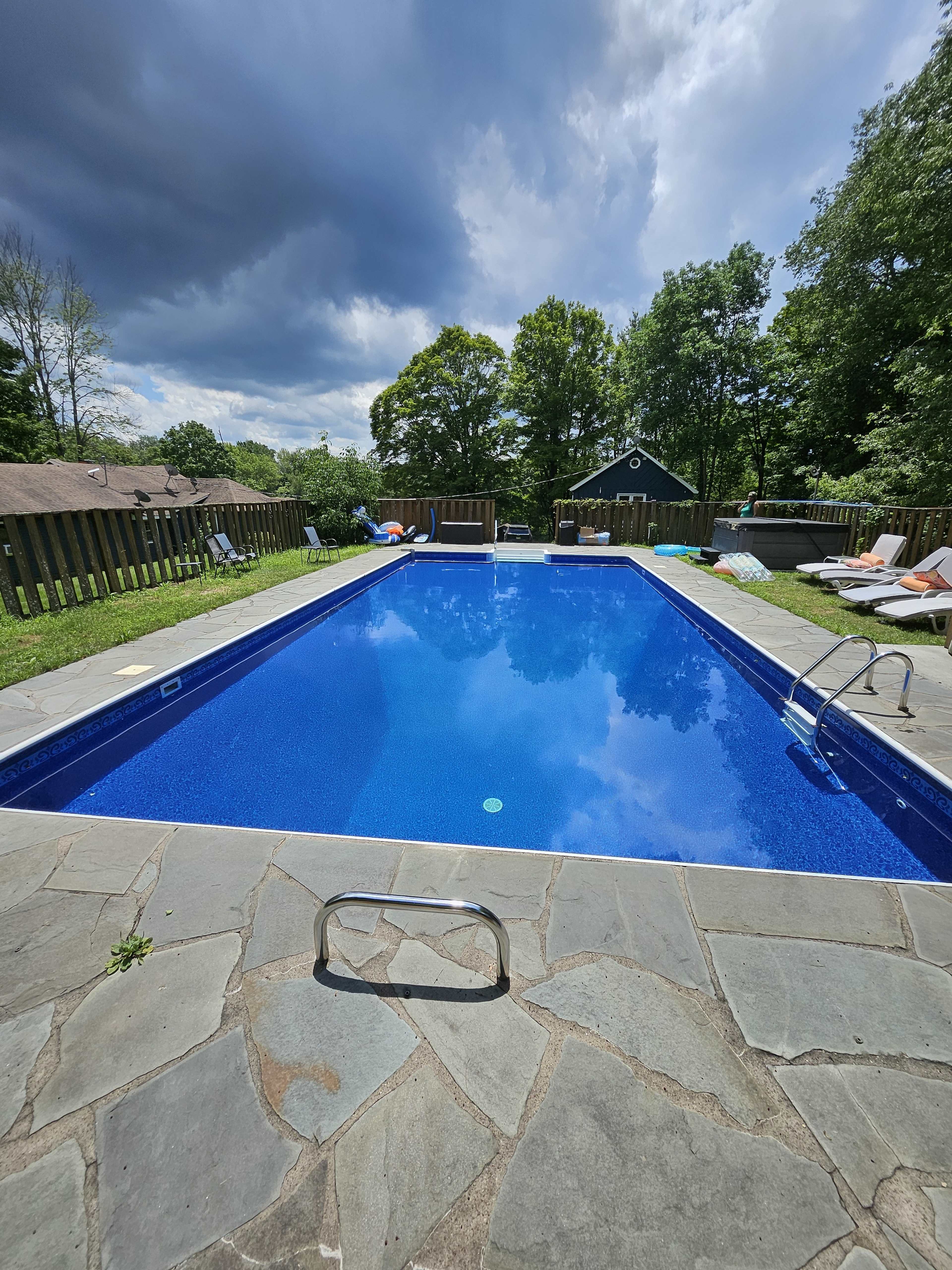 The image shows a rectangular swimming pool surrounded by stone tiles and grassy areas, with houses and trees visible in the background under a cloudy sky.