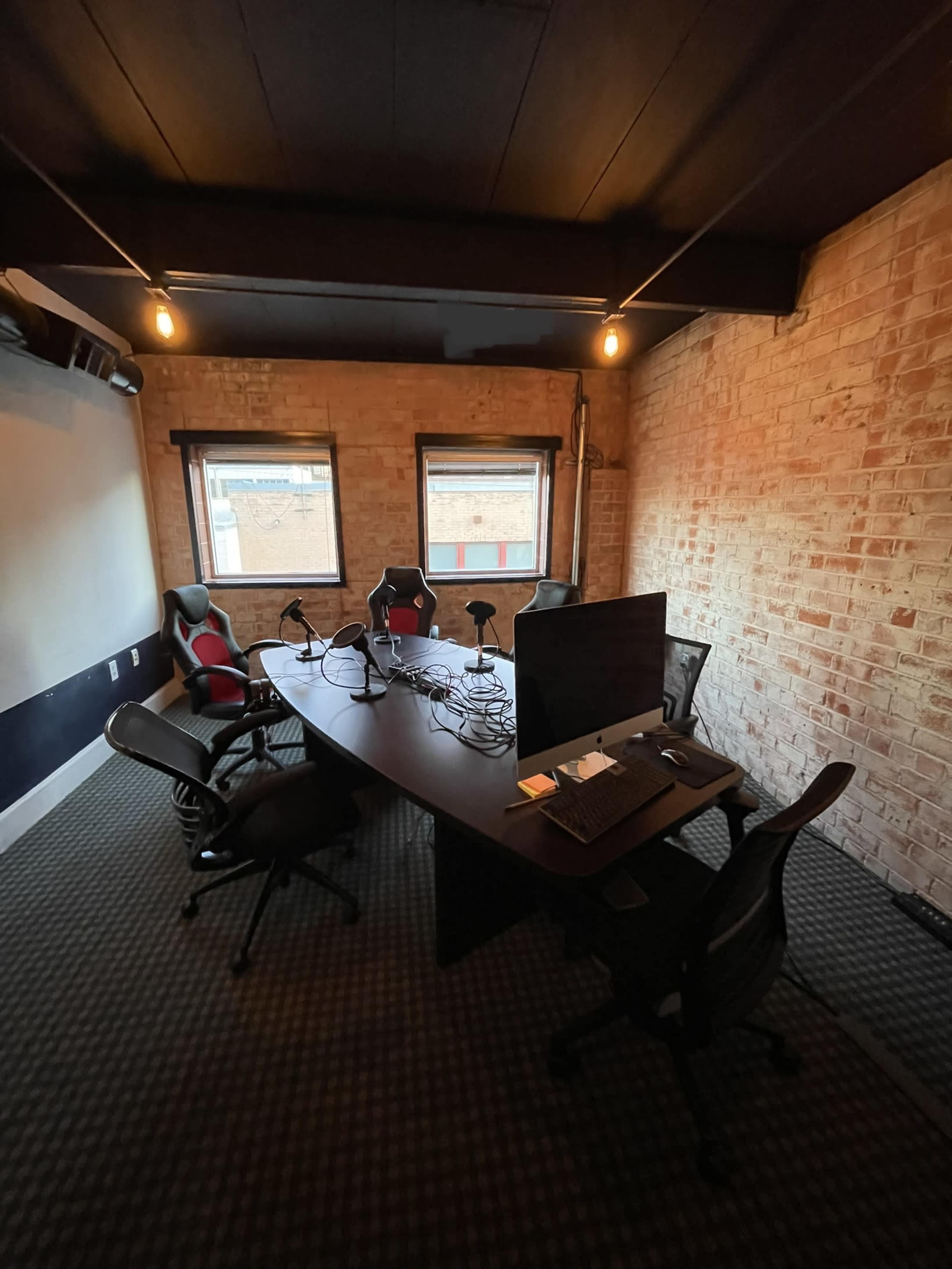 A conference room with a long table surrounded by chairs, featuring microphones and a monitor, and exposed brick walls with two windows.