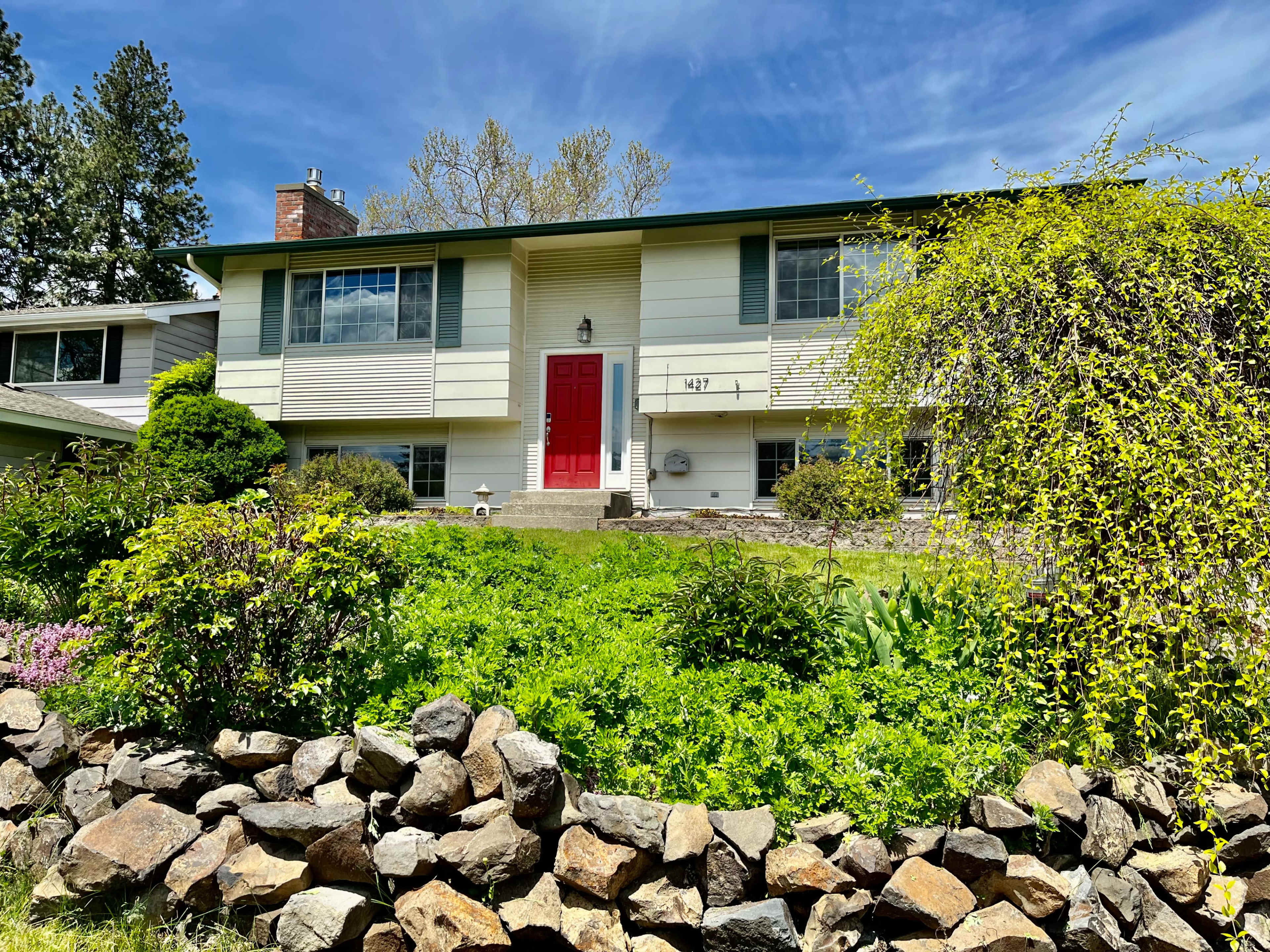 A two-story house with a red front door is surrounded by a well-maintained garden and a rocky border.