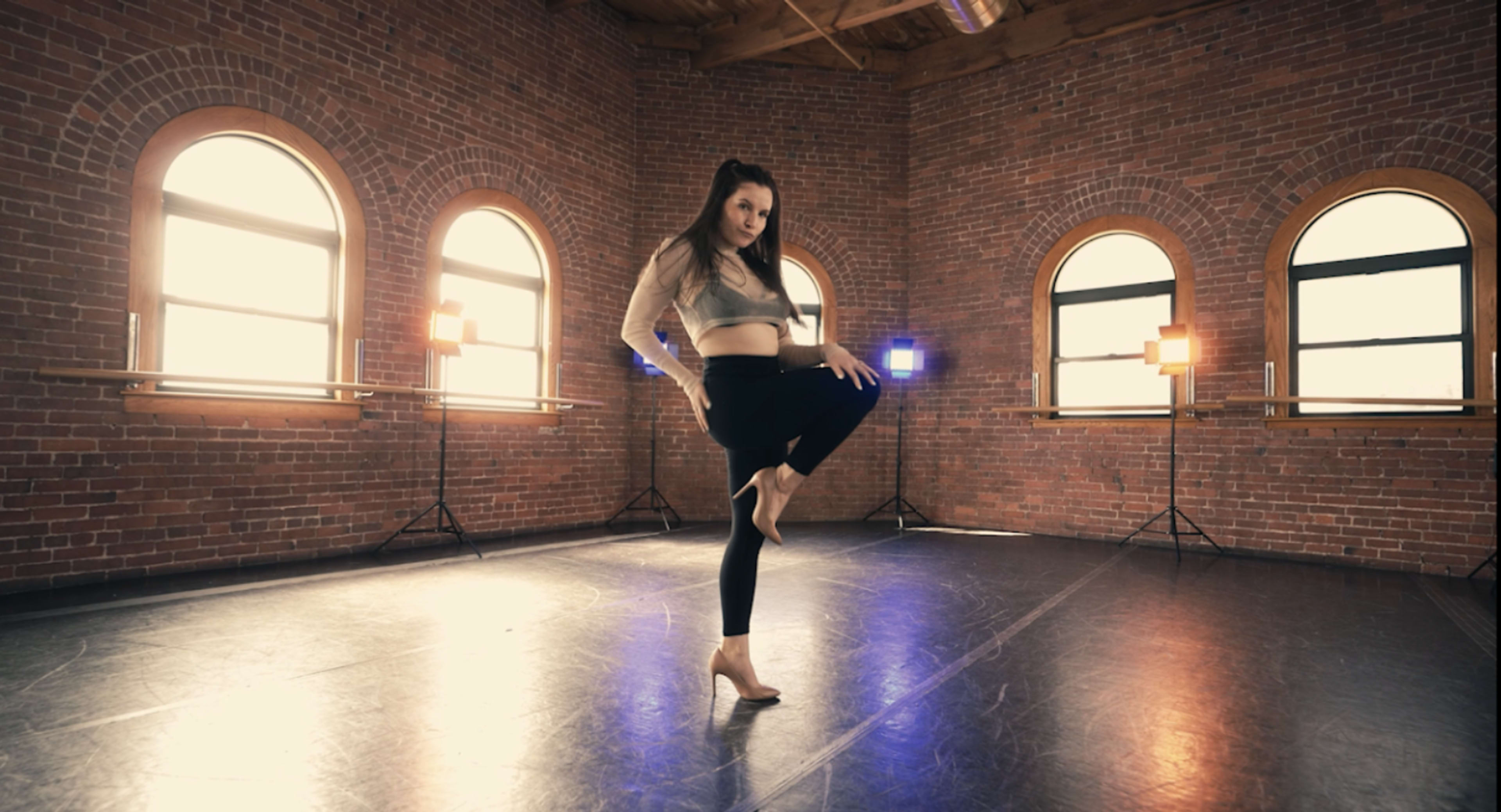 A dancer poses on one leg in a sunlit studio with brick walls and arched windows.