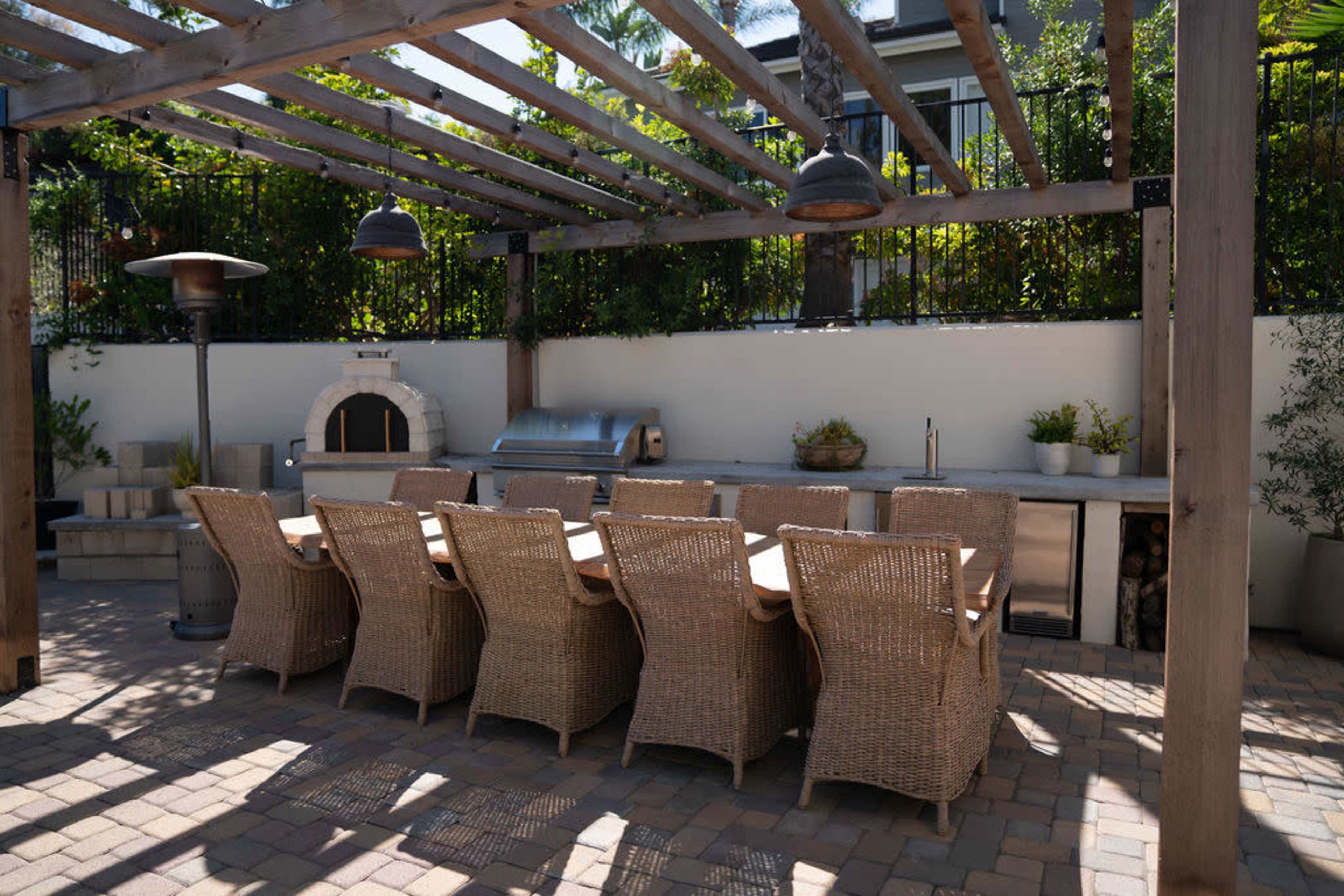 An outdoor dining area with a long wooden table surrounded by wicker chairs, a built-in grill, and a pizza oven under a pergola.