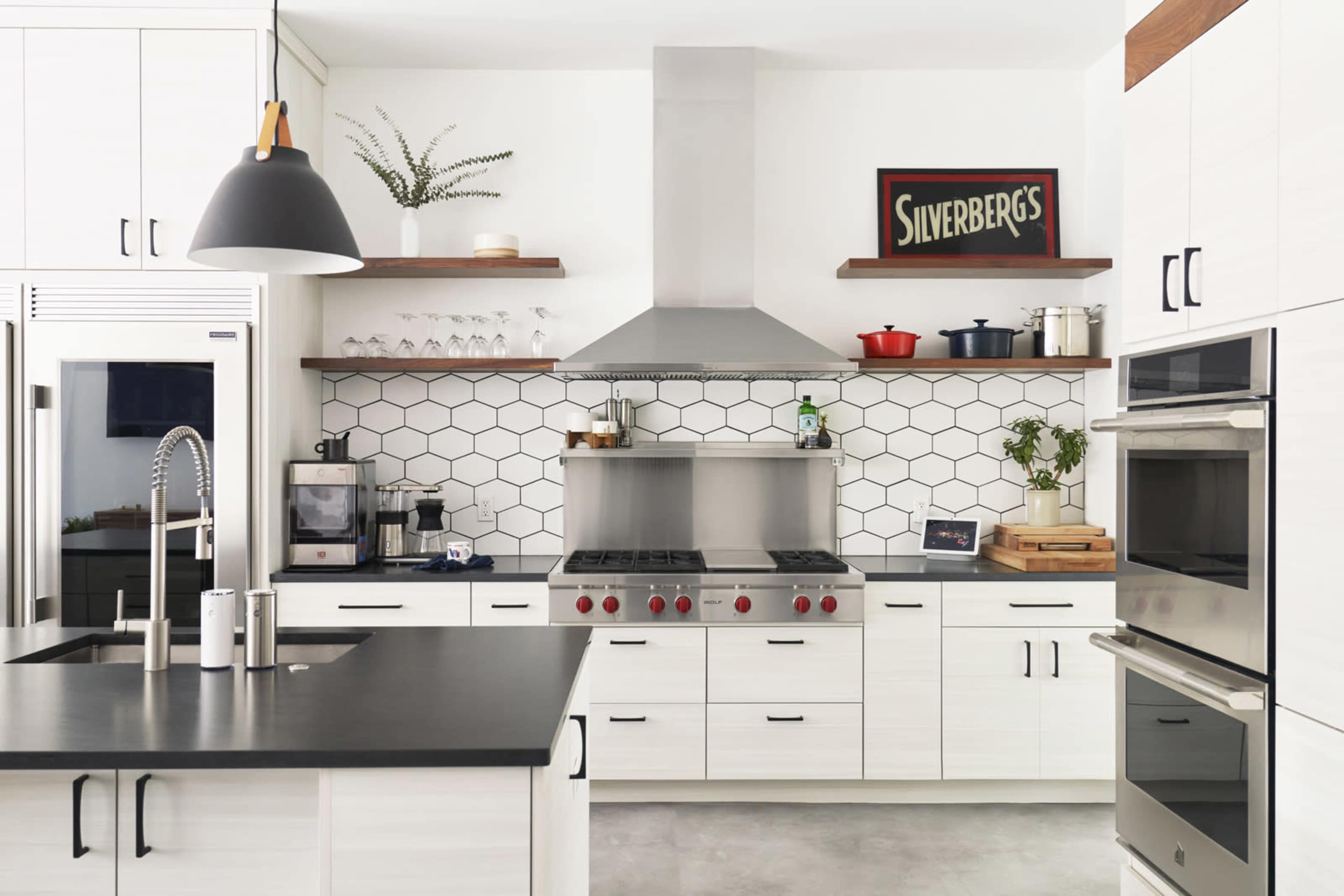 A modern kitchen features white cabinetry, a hexagonal tile backsplash, metal appliances, and a central cooking area with a gas range and overhead hood.
