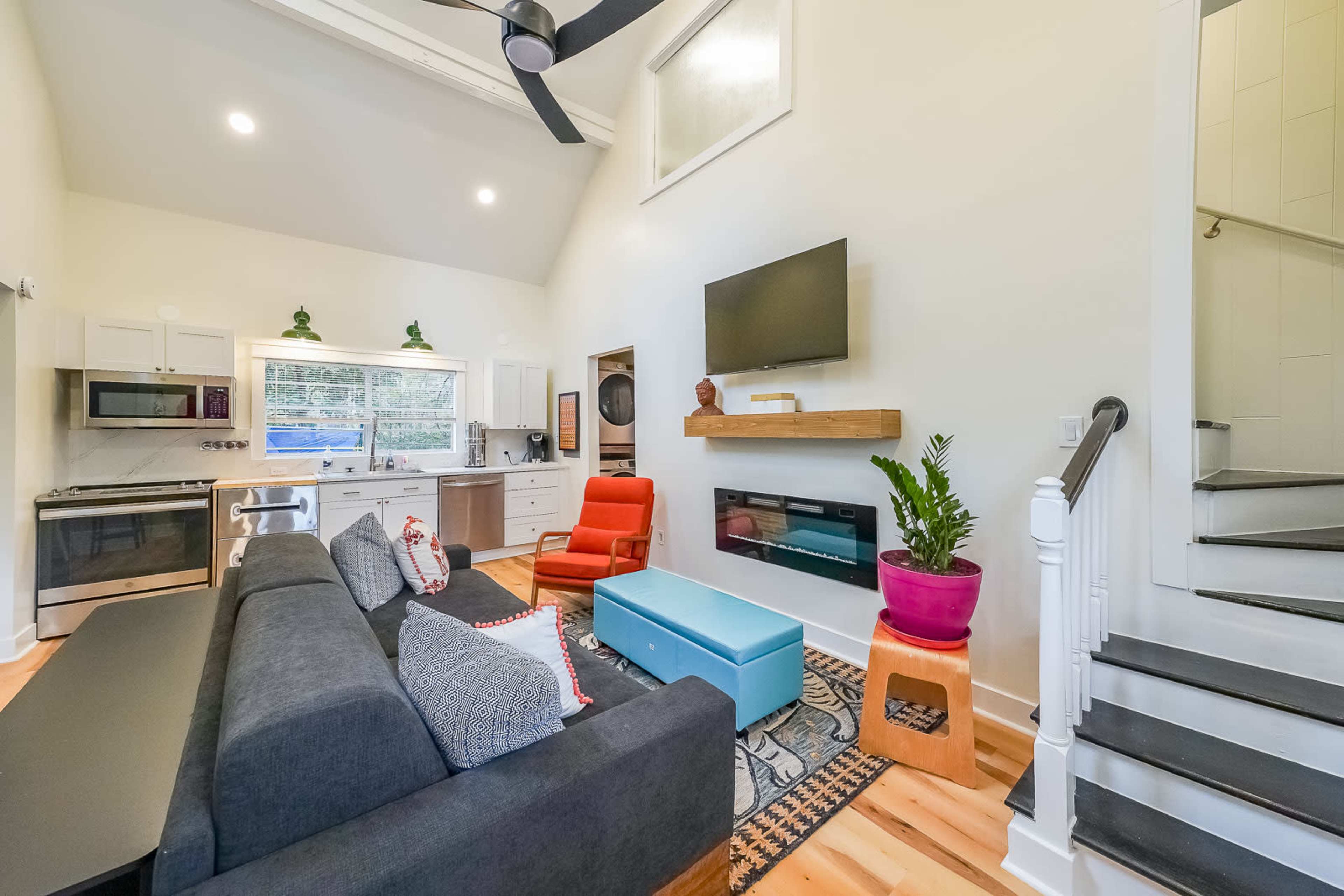 The image shows a modern, open-concept living room with a gray sectional couch, a blue coffee table, and a staircase leading to a loft.