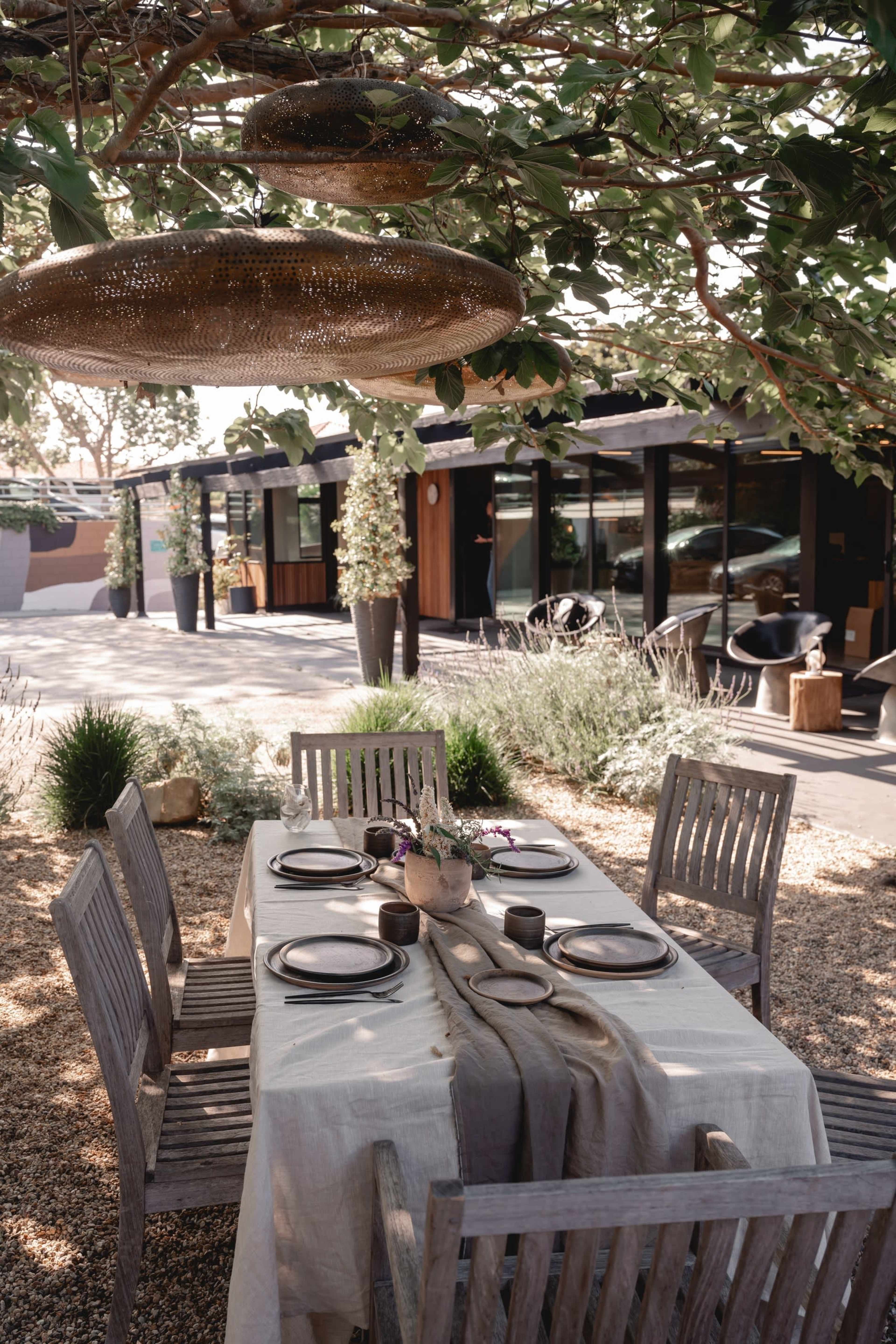 A wooden dining table is set with plates and decorative items under a tree in a patio area adjacent to a modern building.