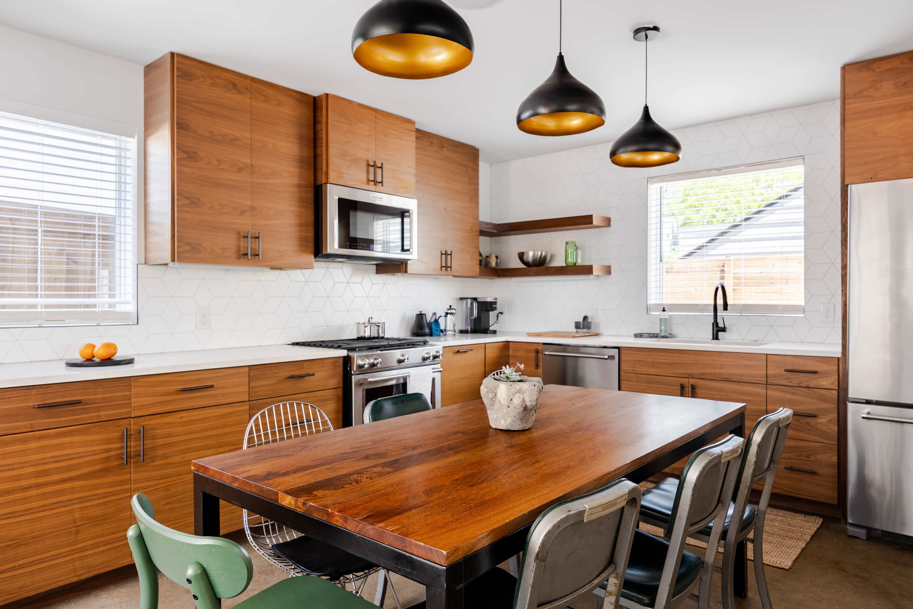 A modern kitchen featuring wooden cabinets, a stainless steel refrigerator, a central wooden dining table, and three pendant lights hanging above.