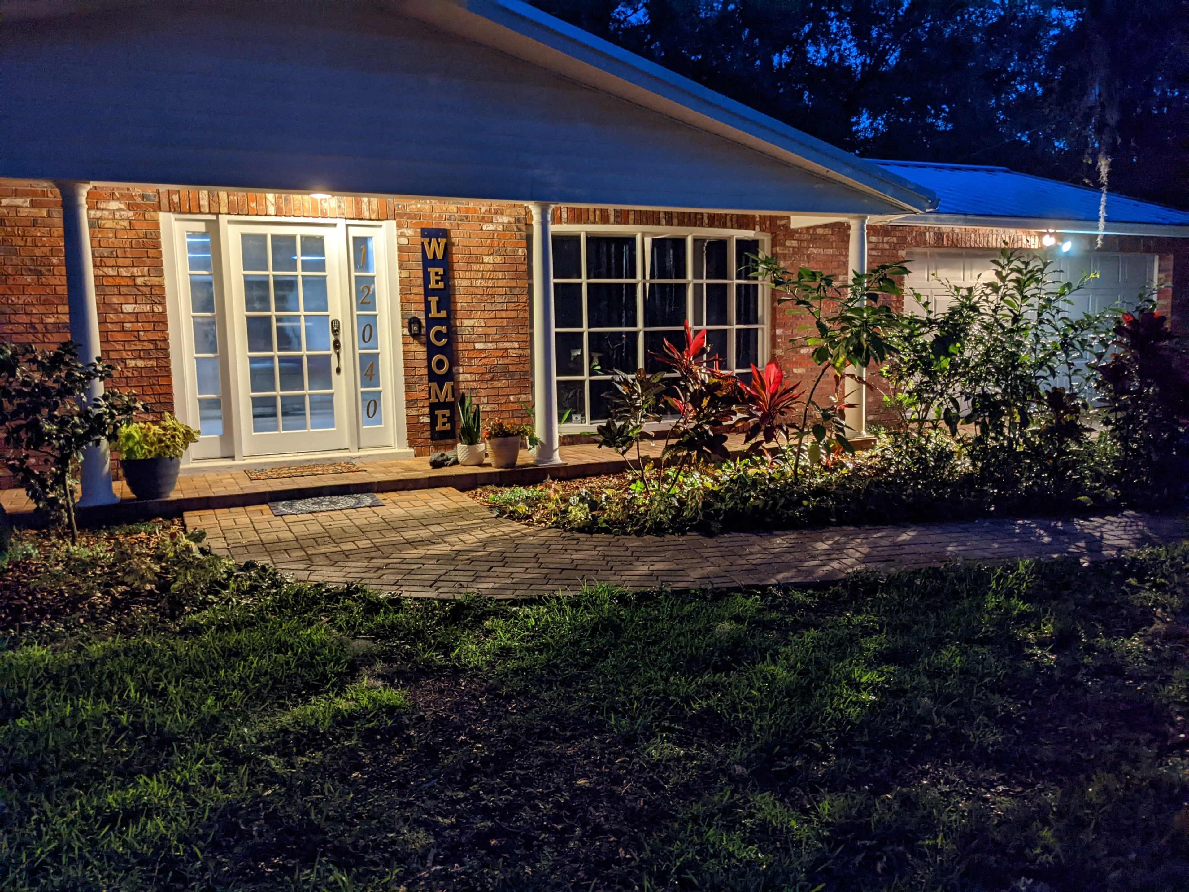 A brick home entrance at dusk, with a "WELCOME" sign displayed next to the front door, surrounded by plants and a walkway made of pavers.