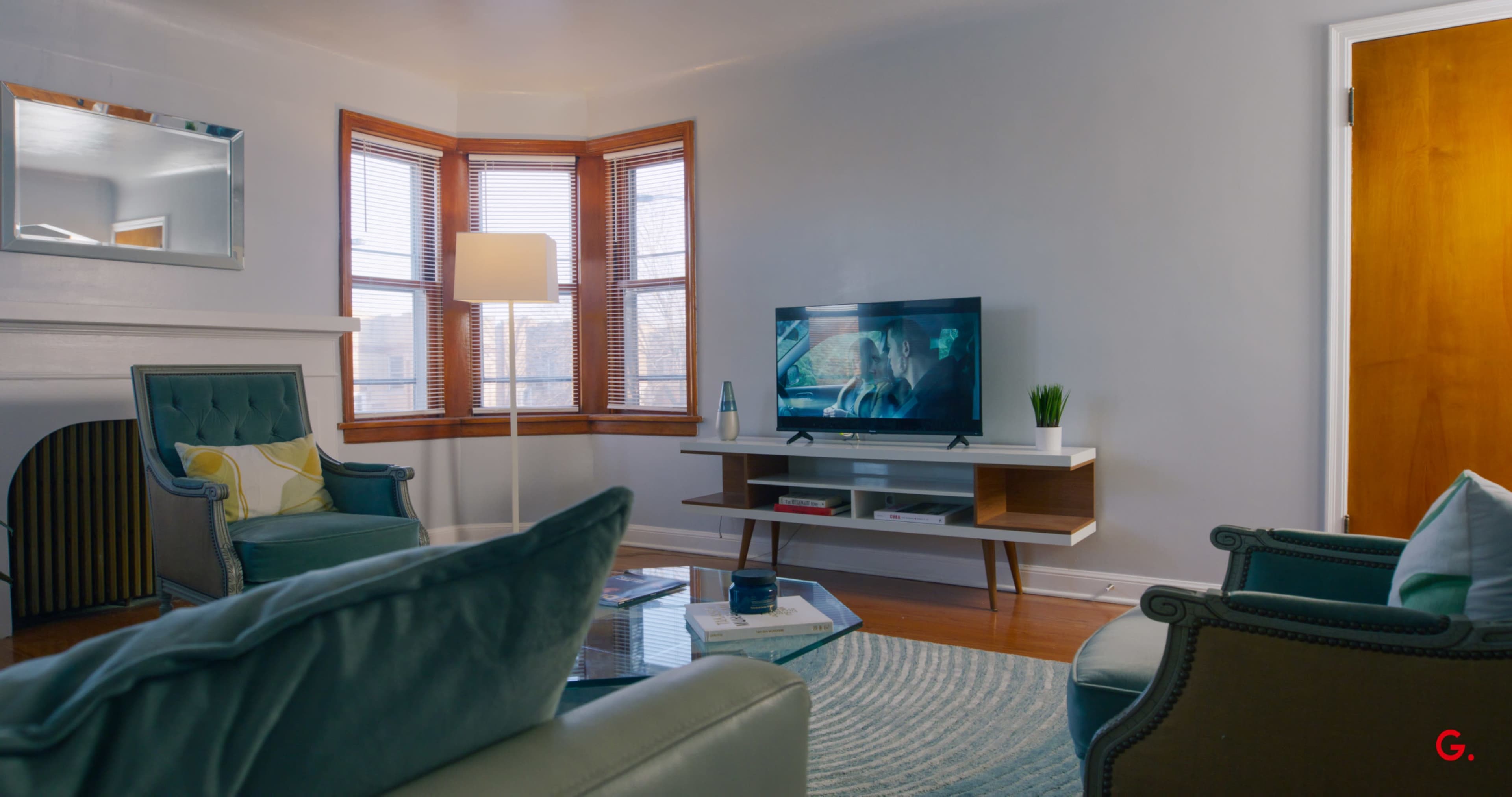 The living room features a television set displaying a movie, a cozy seating arrangement, and large windows with natural light.