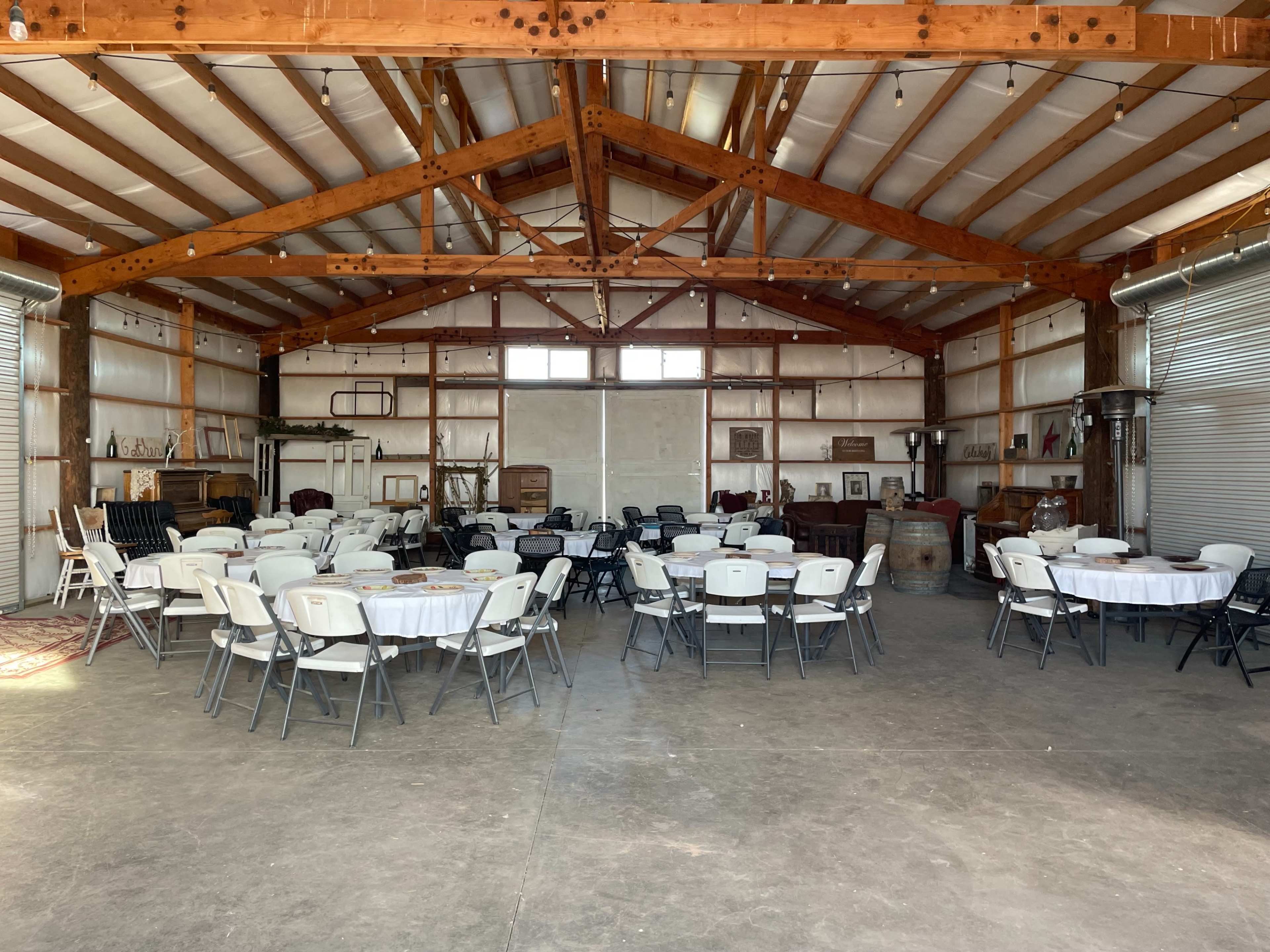 A spacious barn interior features round tables set with white tablecloths, surrounded by chairs, under a wooden beam ceiling.