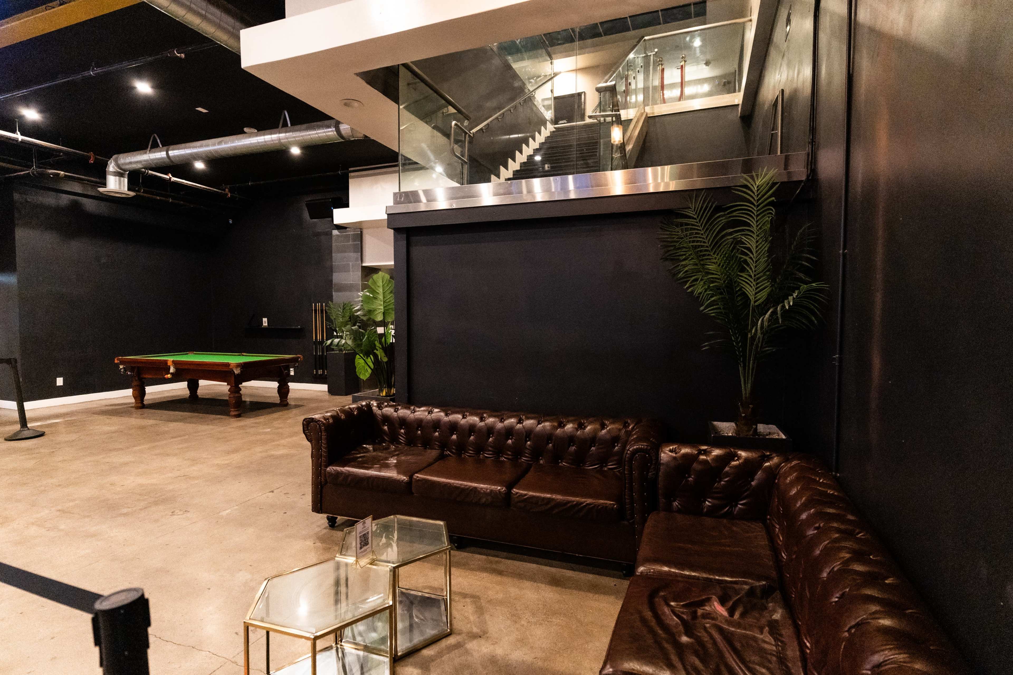 A lounge area with dark walls featuring leather sofas, a glass coffee table, and a pool table in the background beneath a staircase.