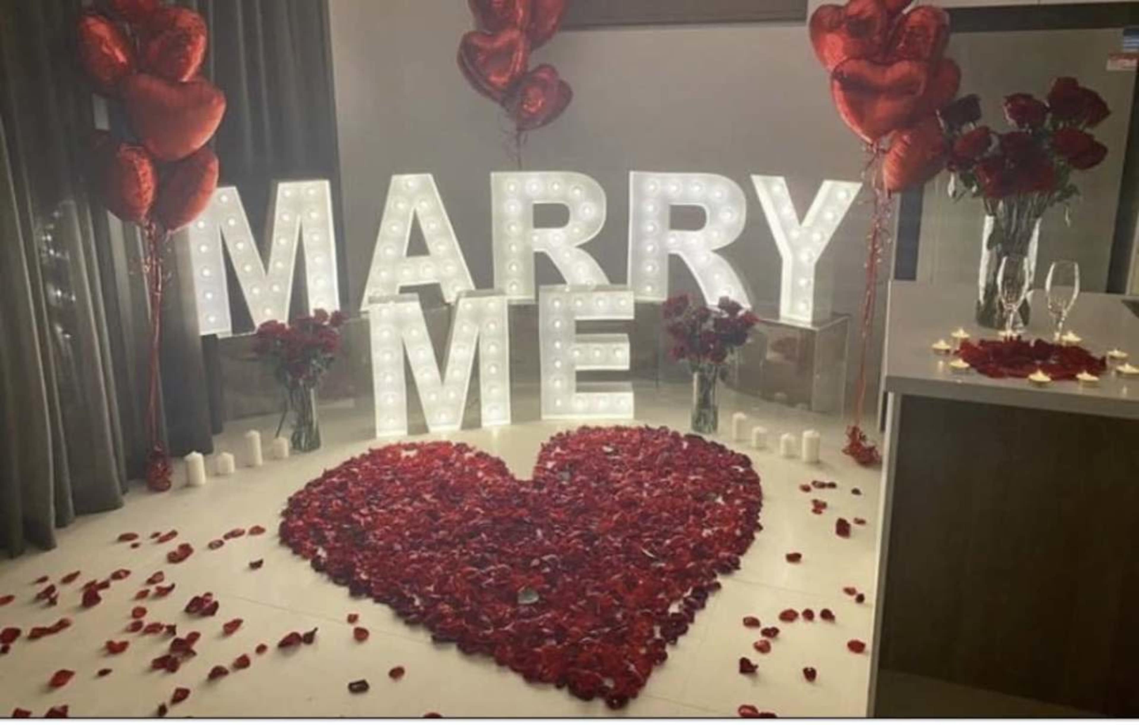 The scene features large illuminated letters spelling "MARRY ME," surrounded by rose petals arranged in a heart shape and decorated with red balloons and candles.