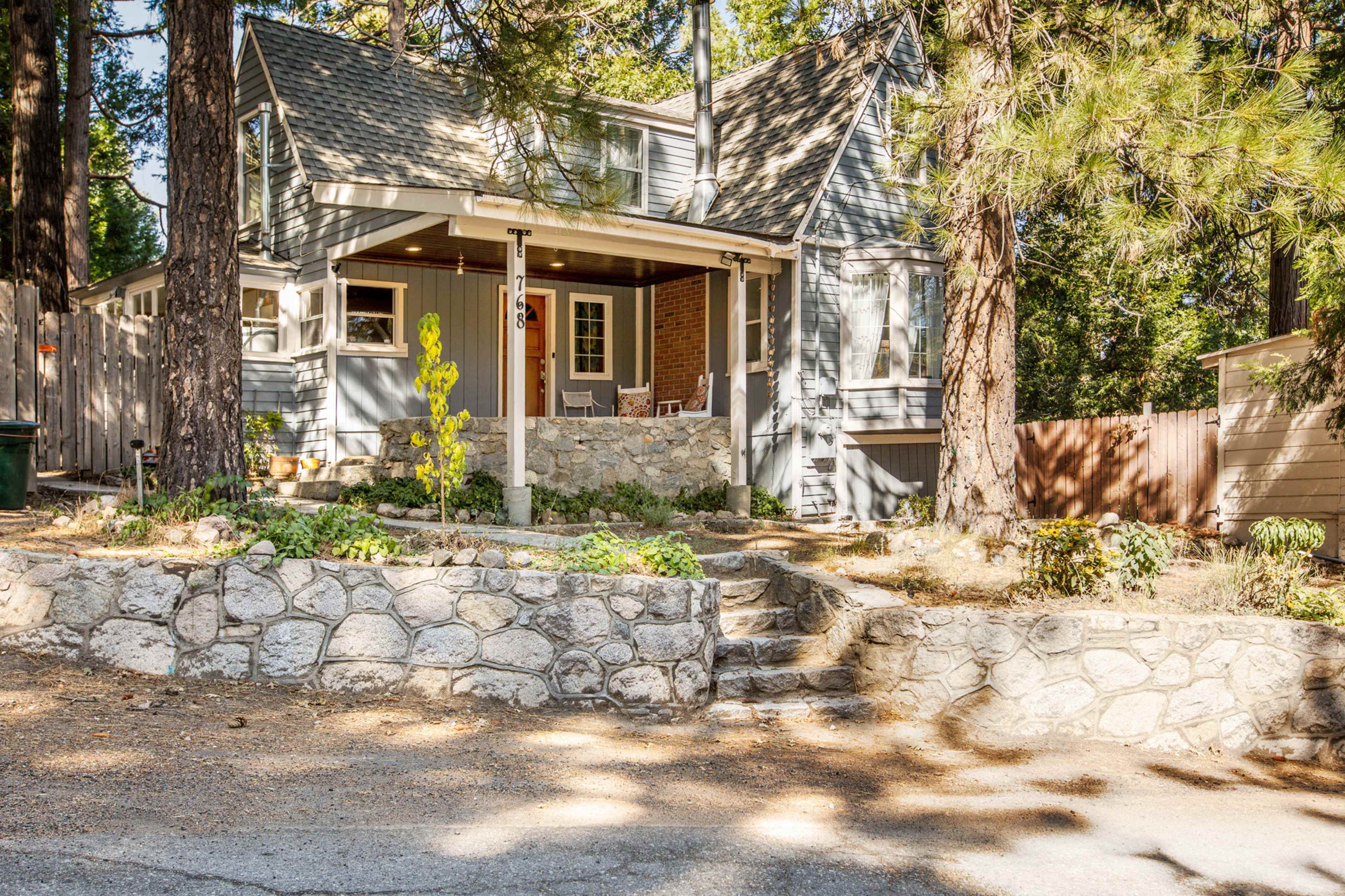 A two-story gray house with a stone porch and surrounded by tall trees and a landscaped yard is visible.