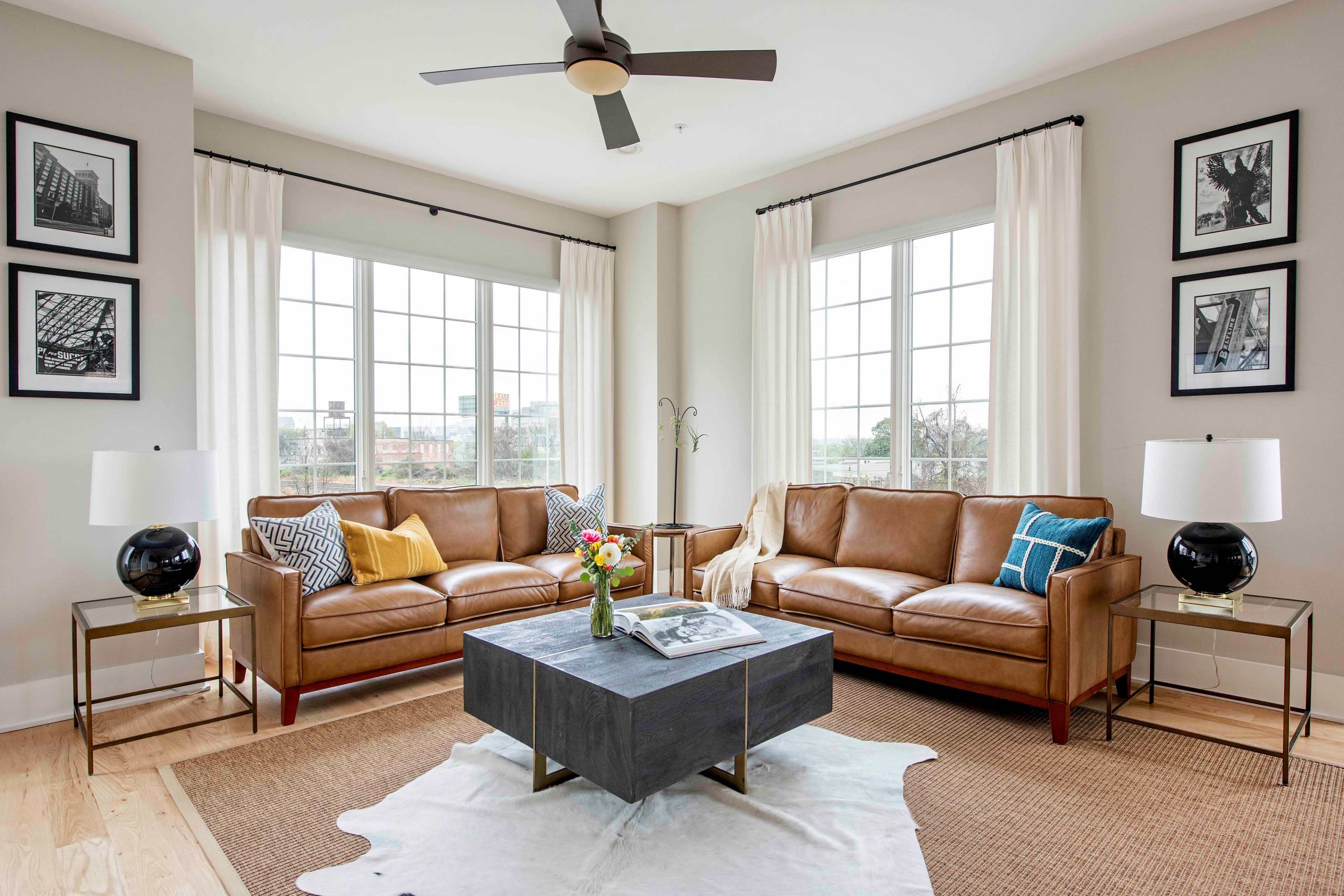 The living room features two brown leather sofas flanking a square coffee table on a light rug, with large windows letting in natural light and framed photographs on the walls.