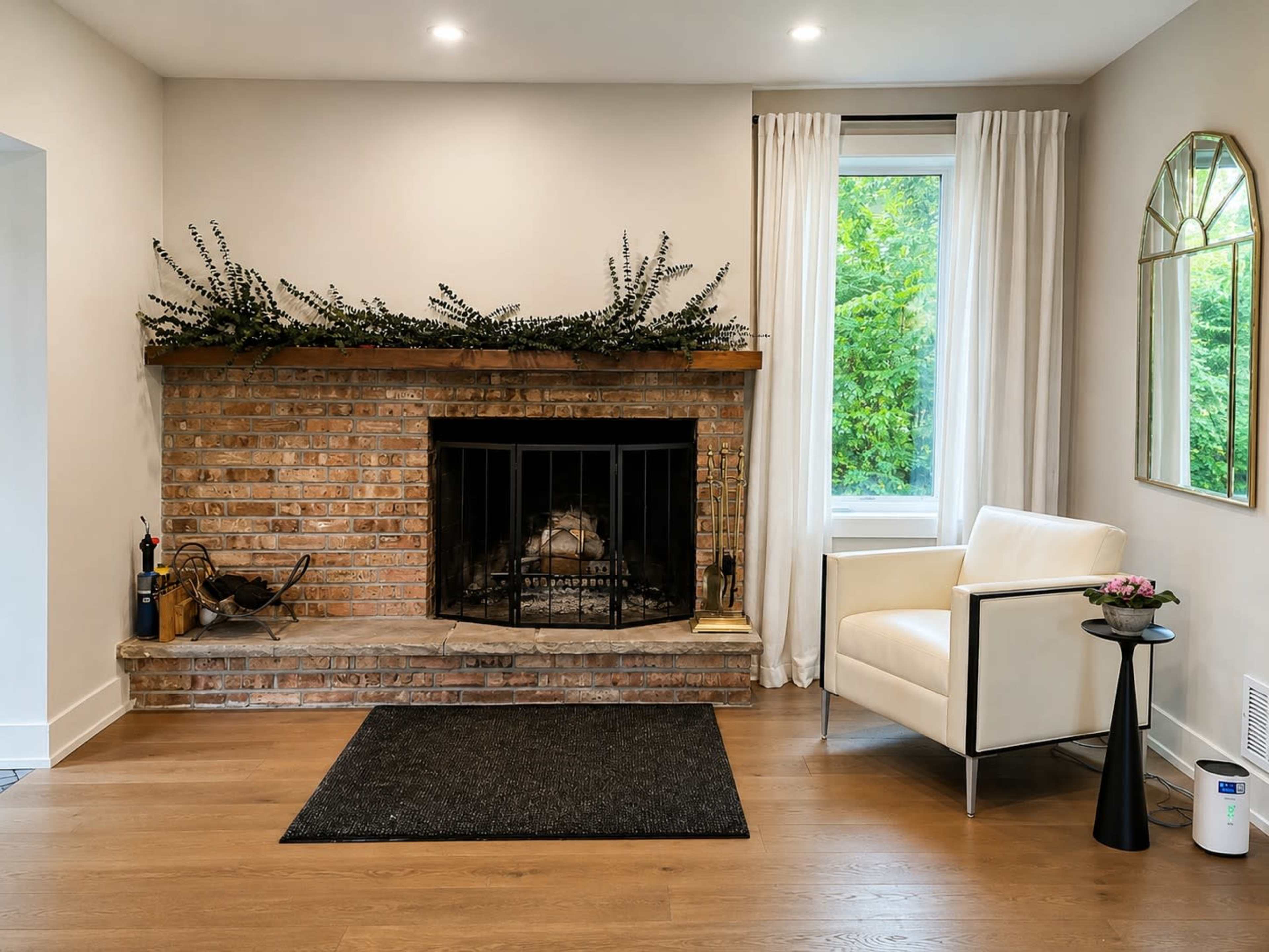 The image shows a modern living room with a brick fireplace, a white armchair, a black side table with a flower arrangement, and large windows draped with light curtains.