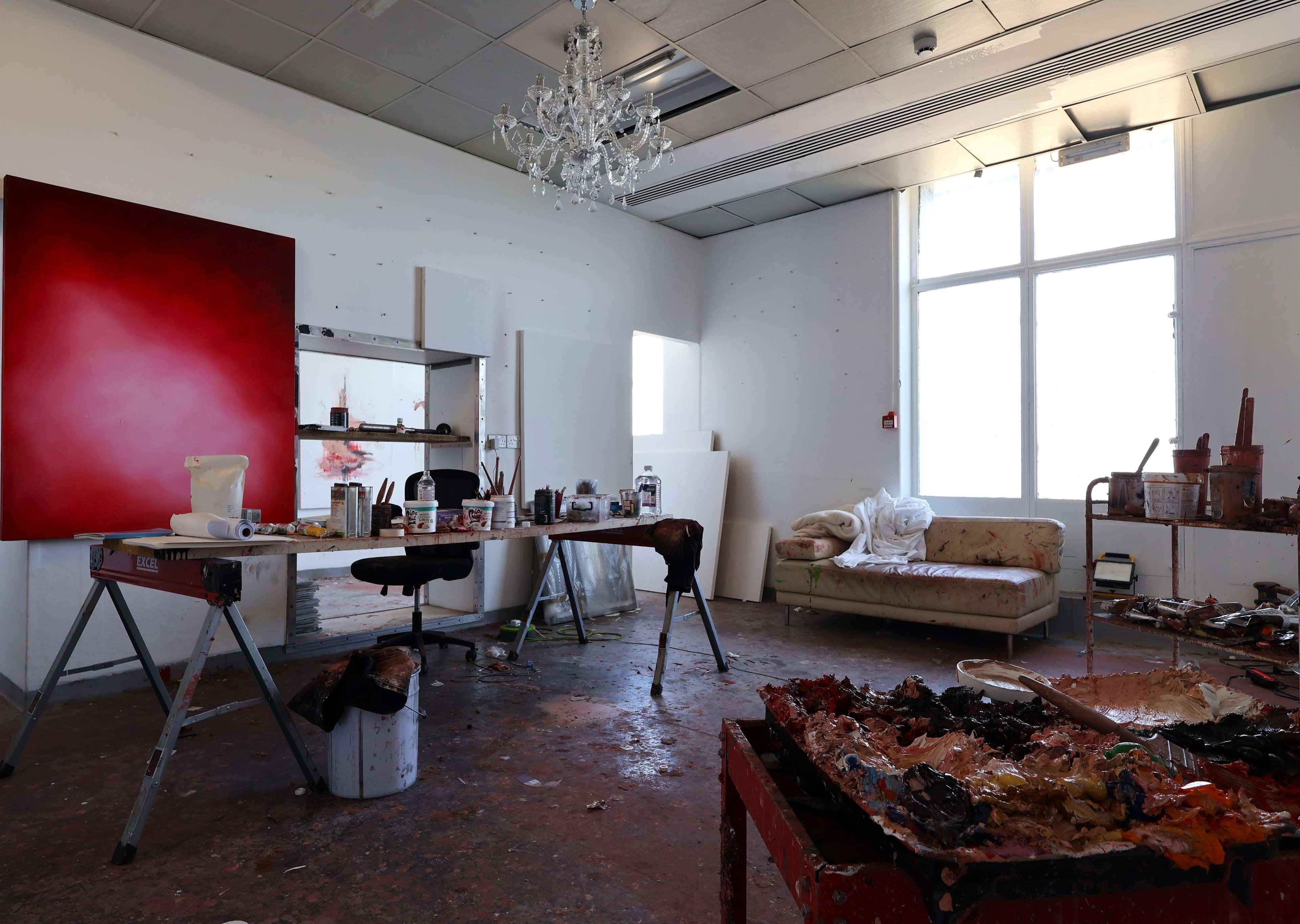 The image shows a spacious artist's studio featuring a large red canvas on an easel, a work table cluttered with painting supplies, and a sofa against the wall.