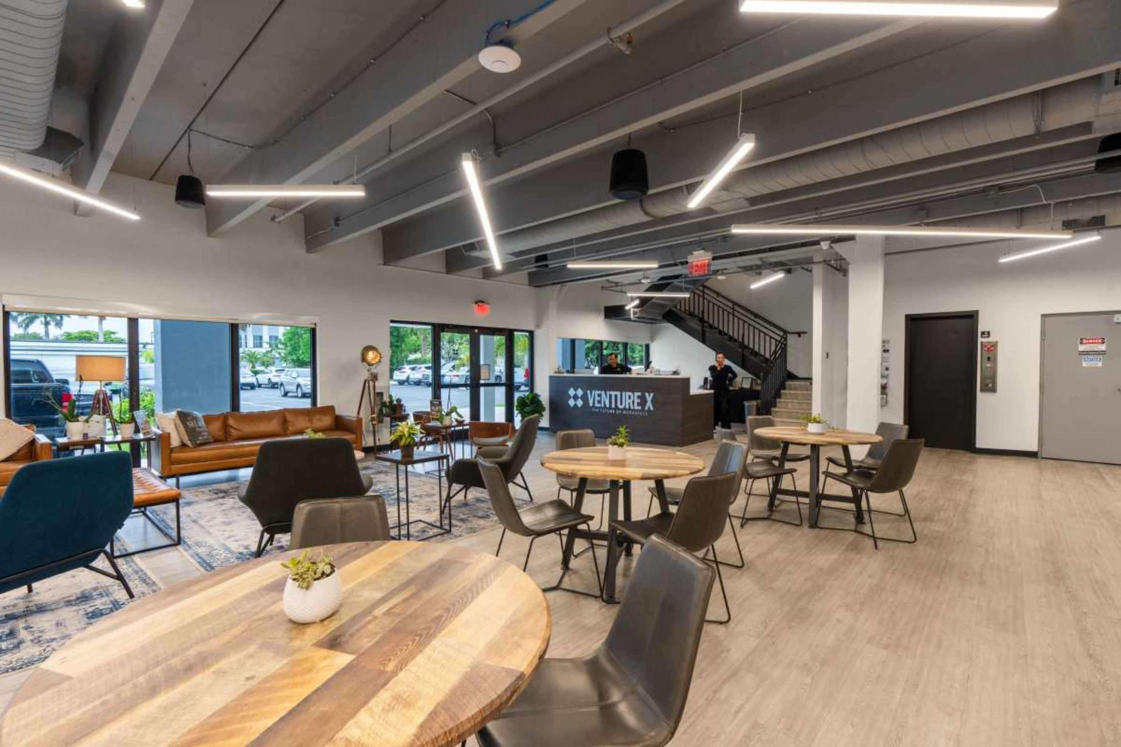 The image shows a modern coworking space featuring wooden tables, chairs, and a reception area with a sign for "Venture X."