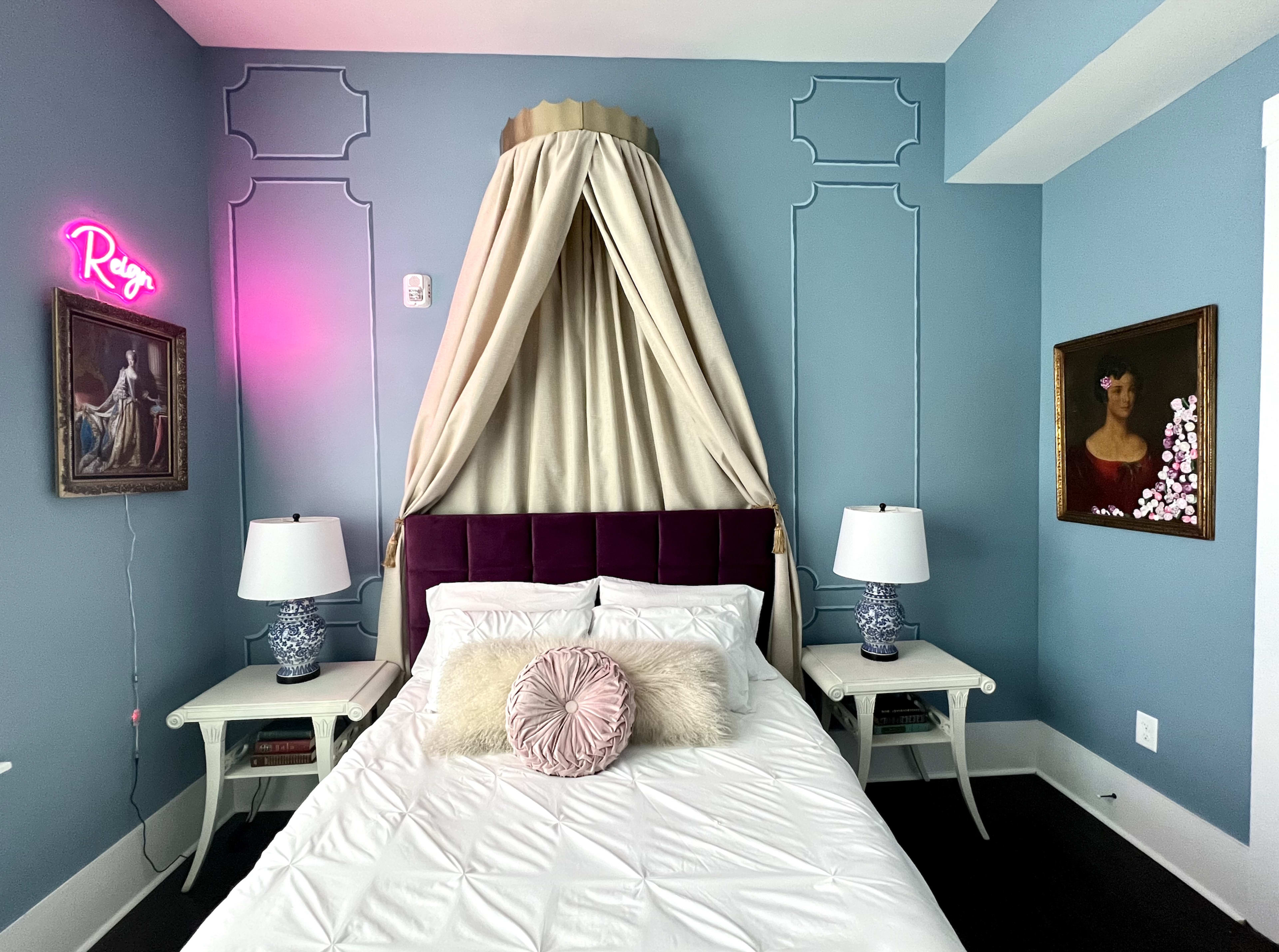 A bedroom featuring a bed with a canopy, two bedside tables with lamps, and decorated walls in a light blue color with artwork and a neon sign.