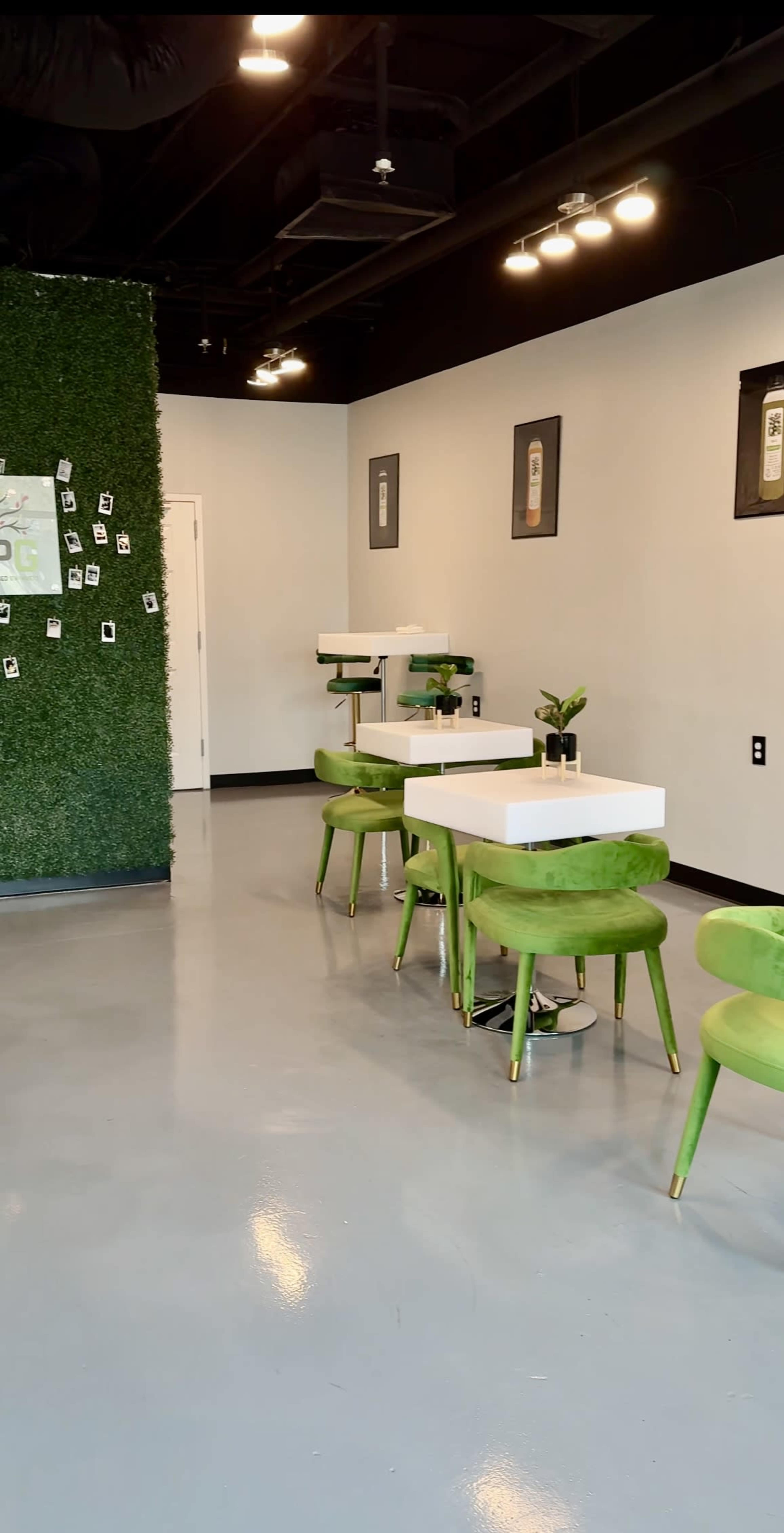 The image shows a modern cafe interior with green chairs, white tables, and a grass-covered wall decorated with polaroid photos.