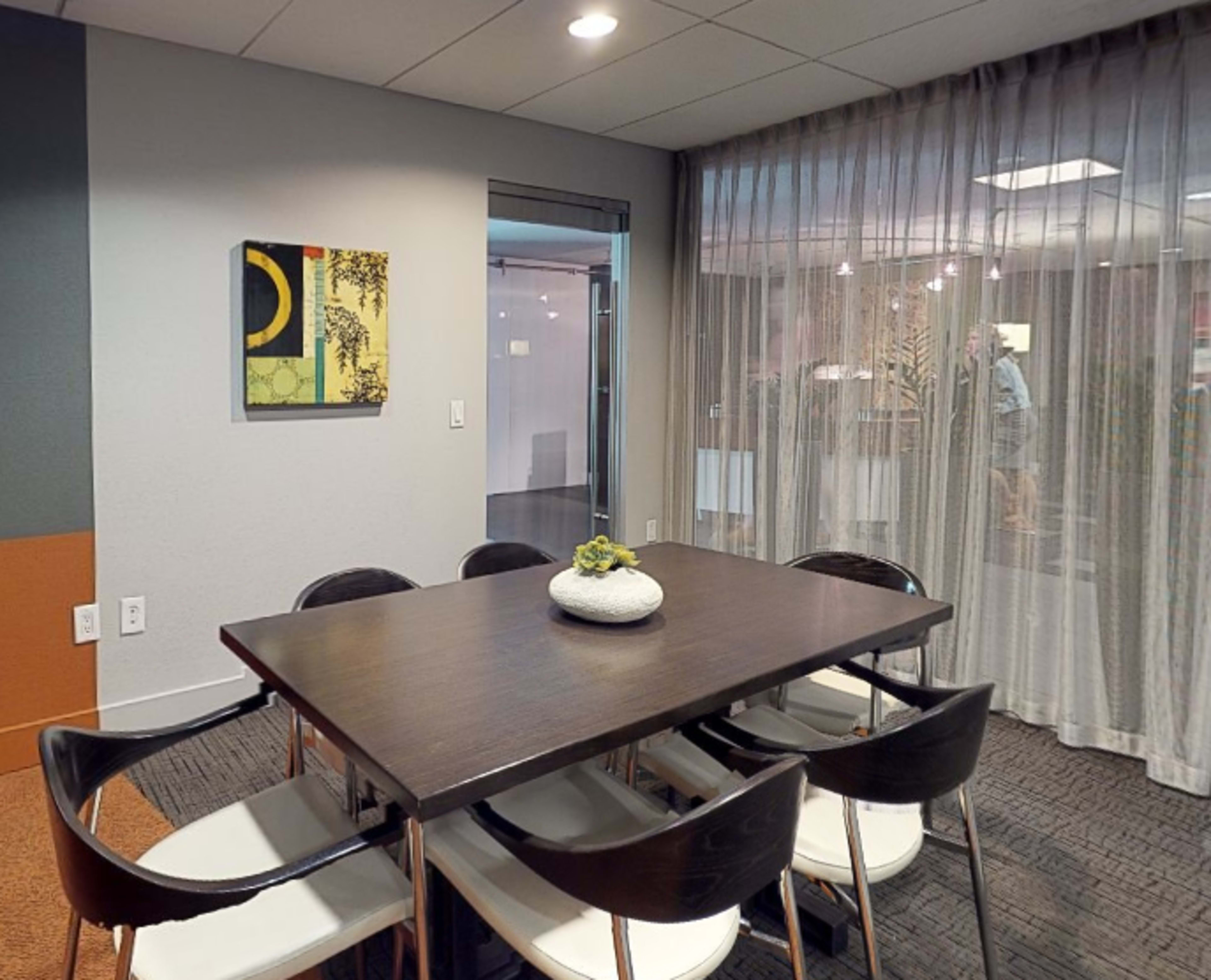 A modern conference room features a dark wooden table surrounded by white chairs, with a framed artwork on the wall and sheer curtains covering a glass partition.
