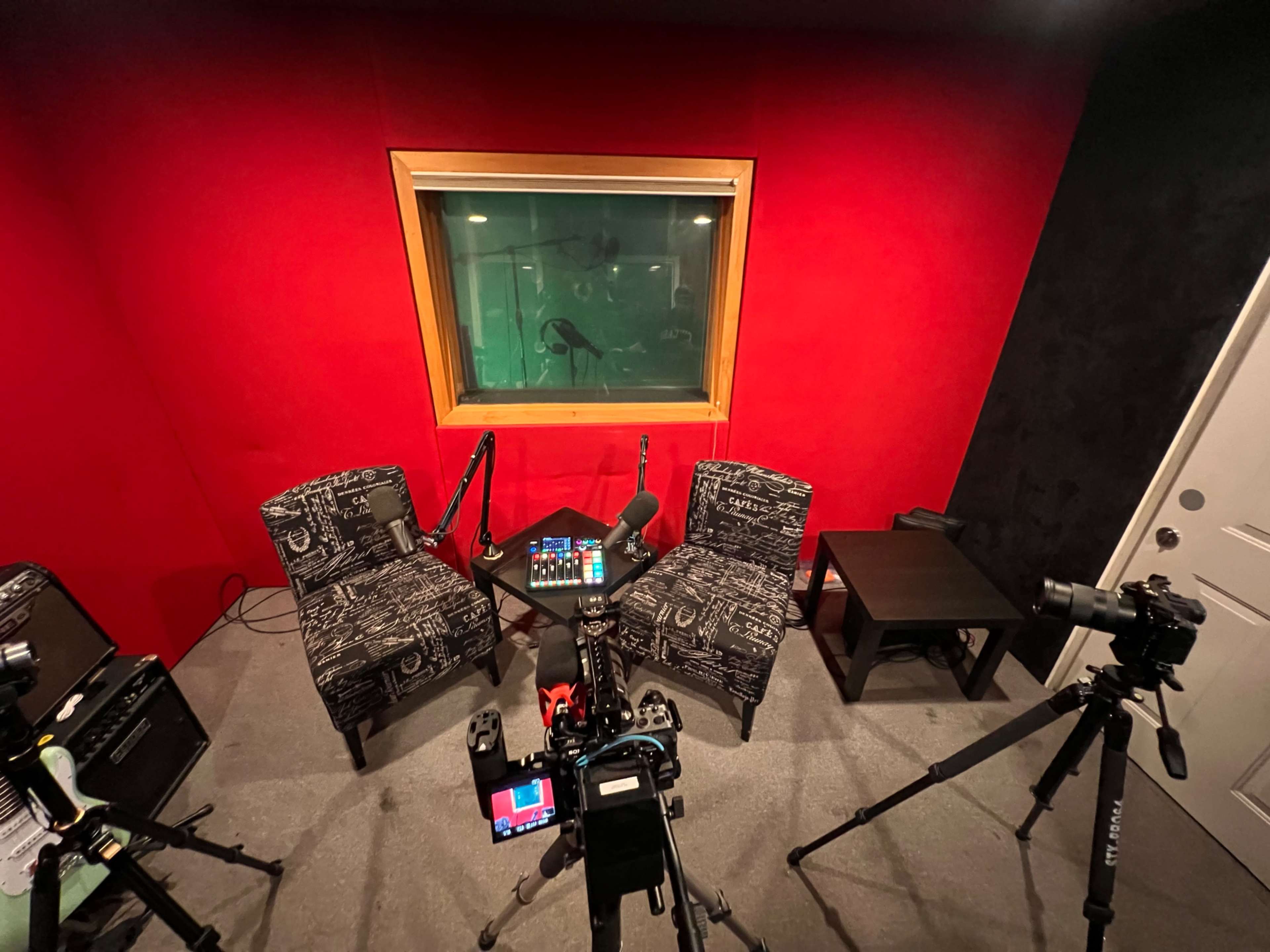 The image shows a recording studio with two chairs, a control panel, and multiple cameras positioned around a glass-walled booth.