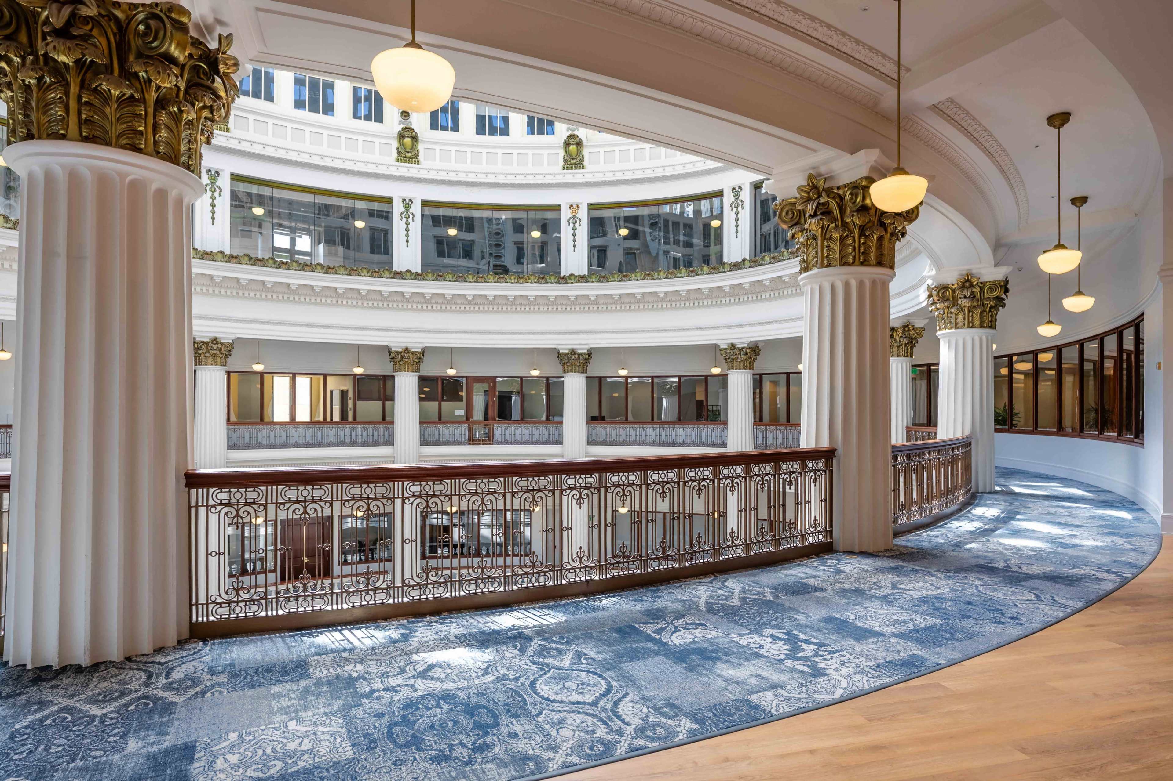 The image shows a spacious, multilevel interior with ornate columns, large windows, and a decorative railing overlooking a central area.