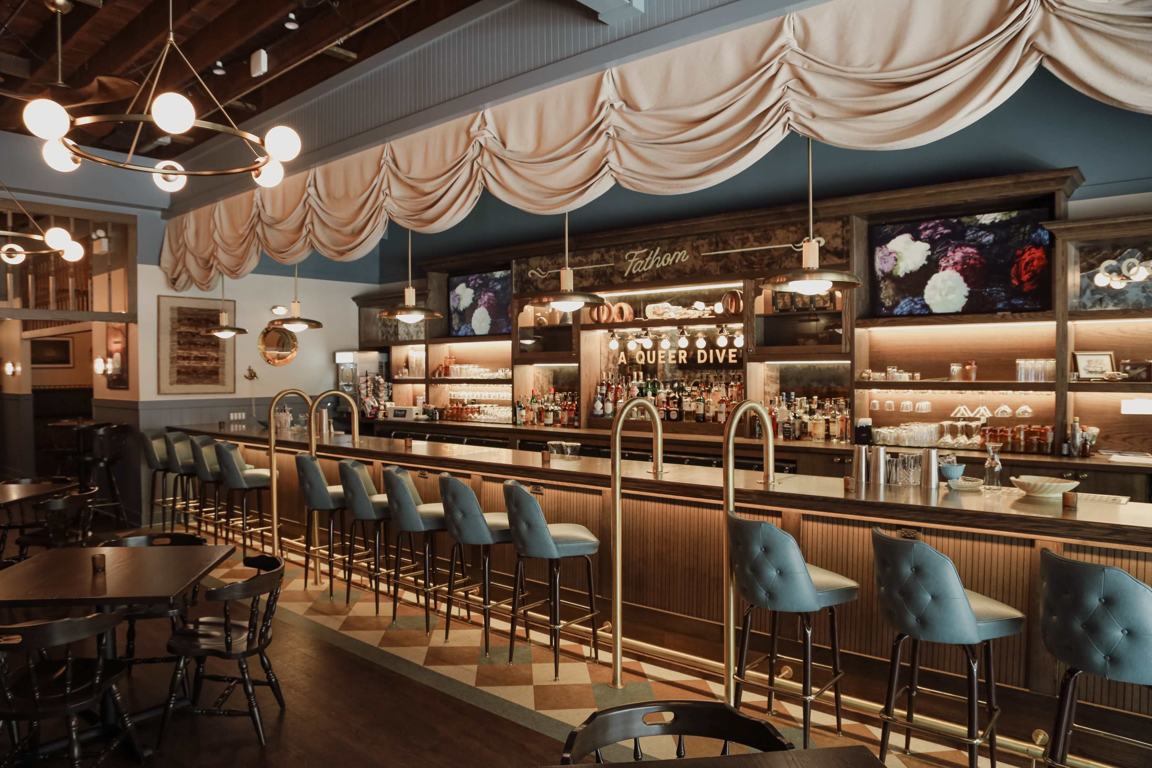 The image shows a stylish bar area with a wooden counter, upholstered blue chairs, and shelves stocked with various bottles, all under elegant draped fabric and soft lighting.