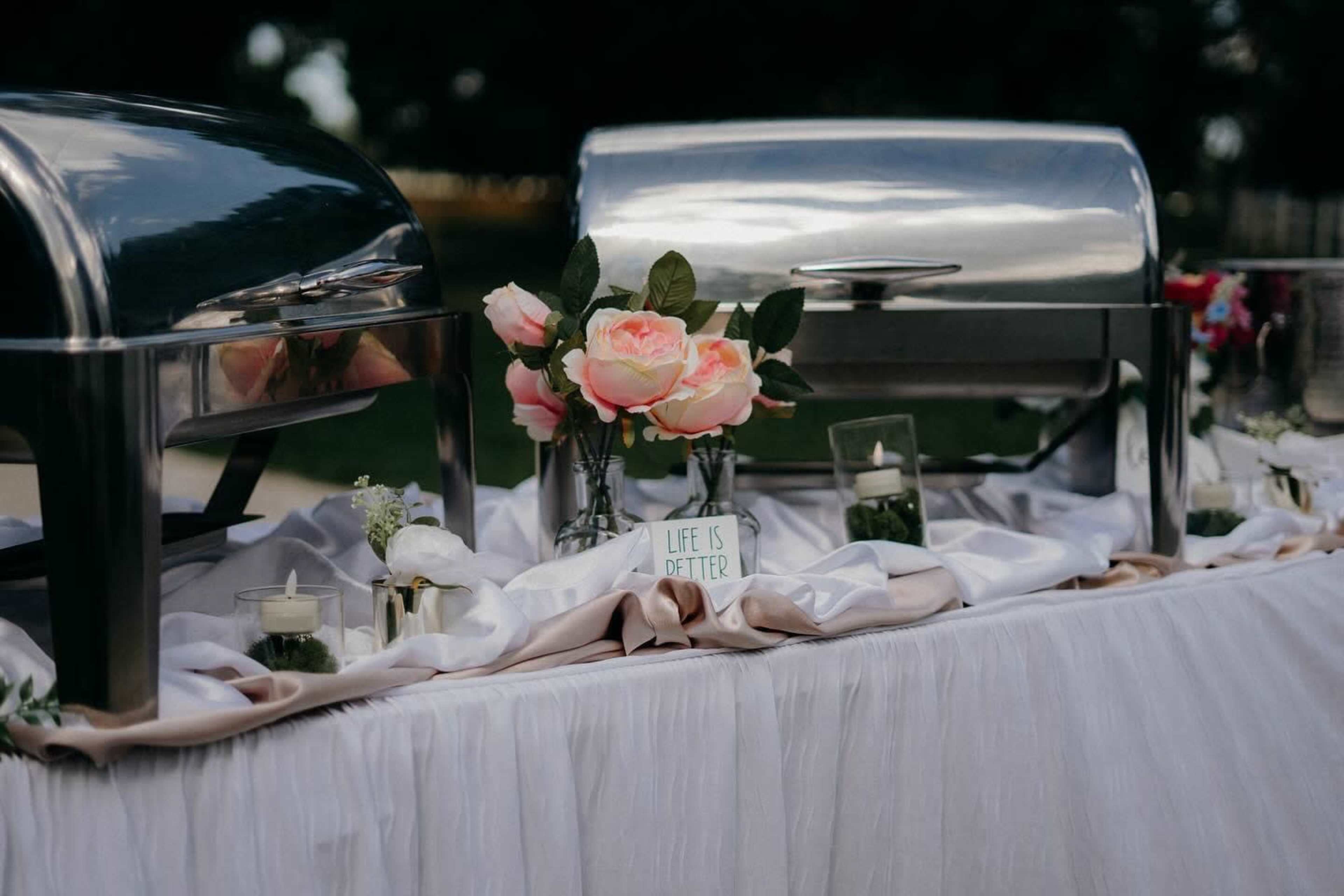 A buffet table is set up with silver food warmers, pink flowers, and a sign that reads "LIFE IS BETTER."