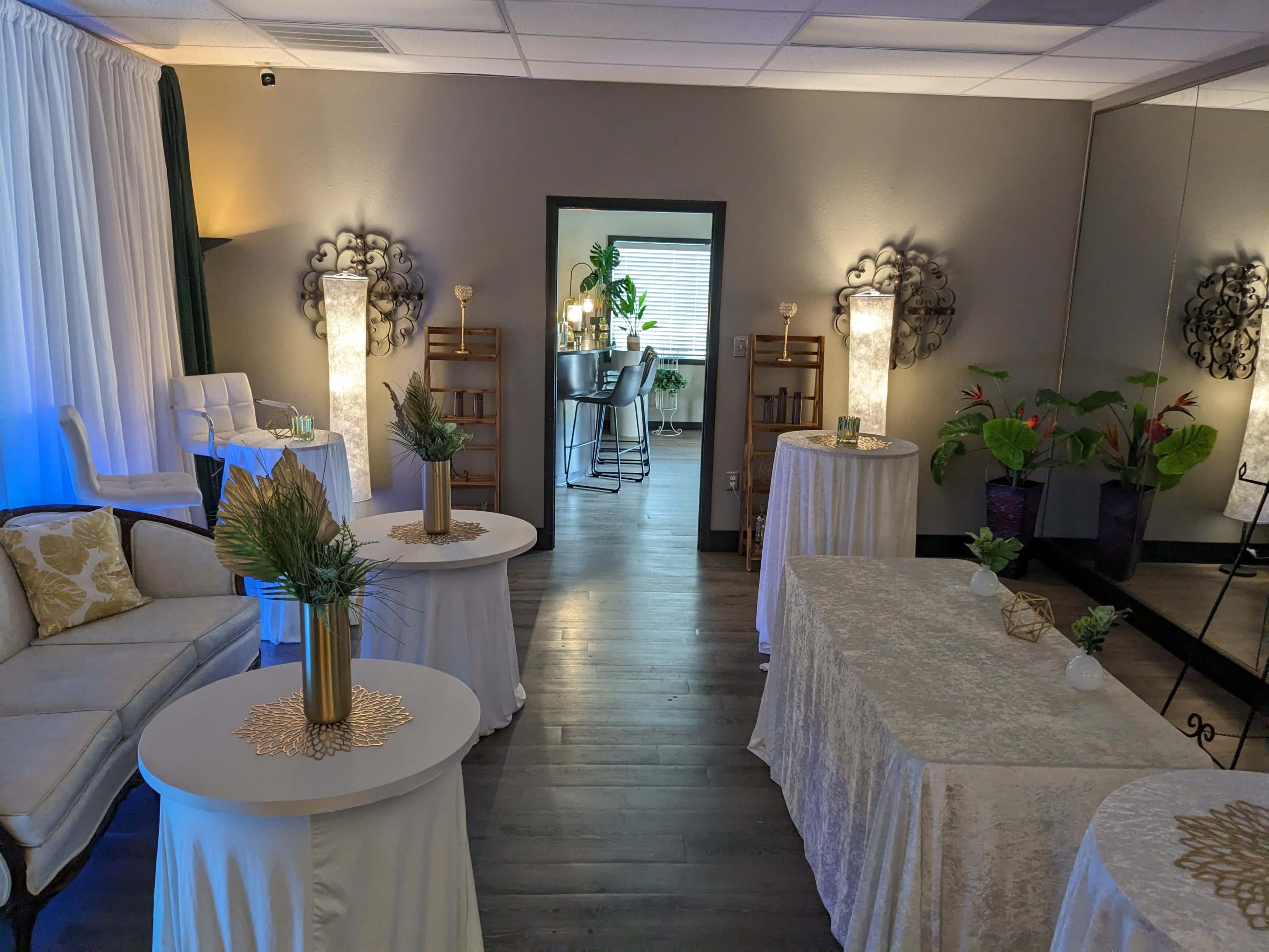 The image shows a cozy room with elegantly arranged tables, a sofa, and decorative plants, leading to a separate area that includes bar stools and shelves.