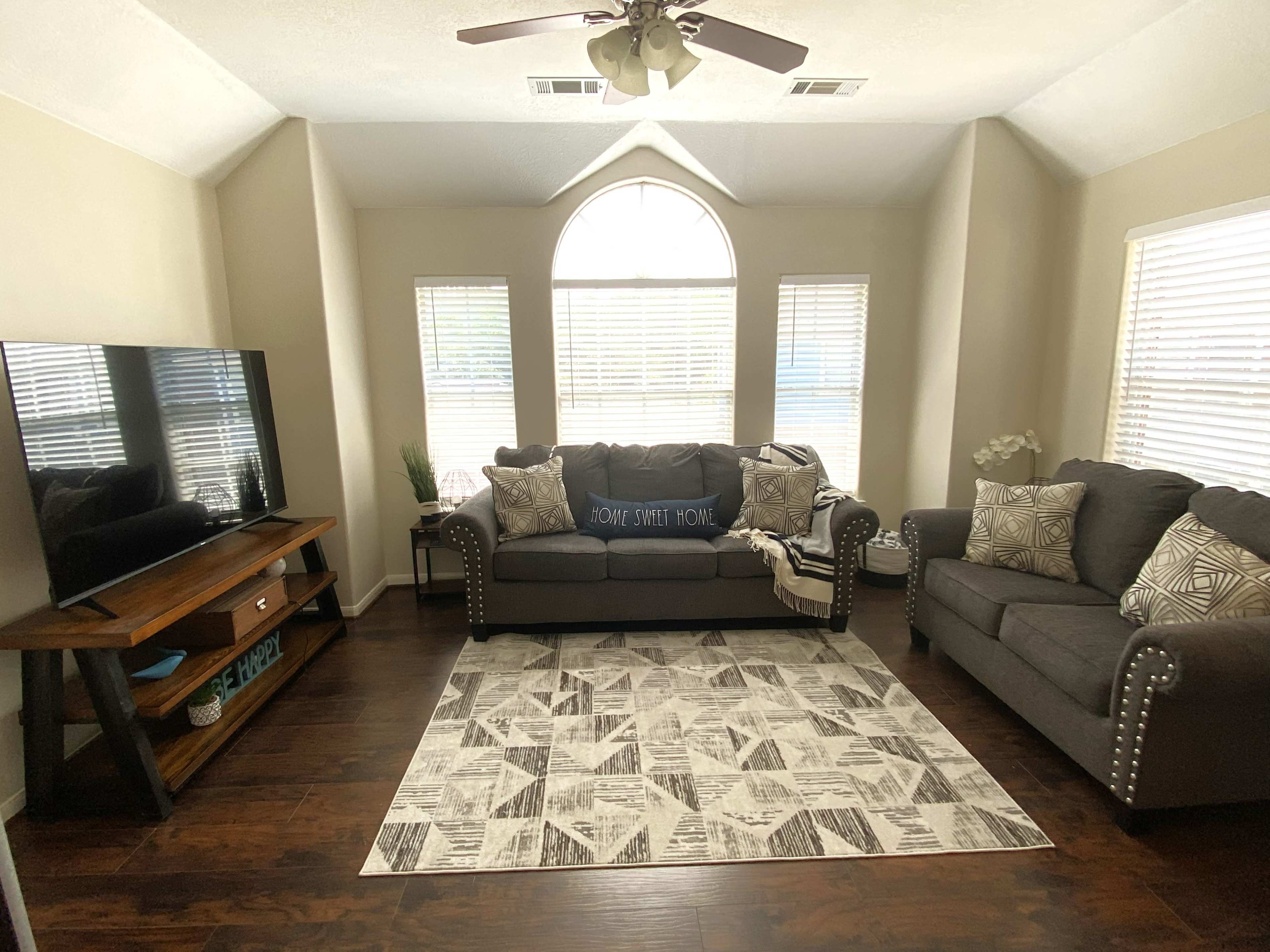 The living room features a large sectional sofa and armchair, a wooden TV stand, and a patterned area rug beneath a bright arched window.