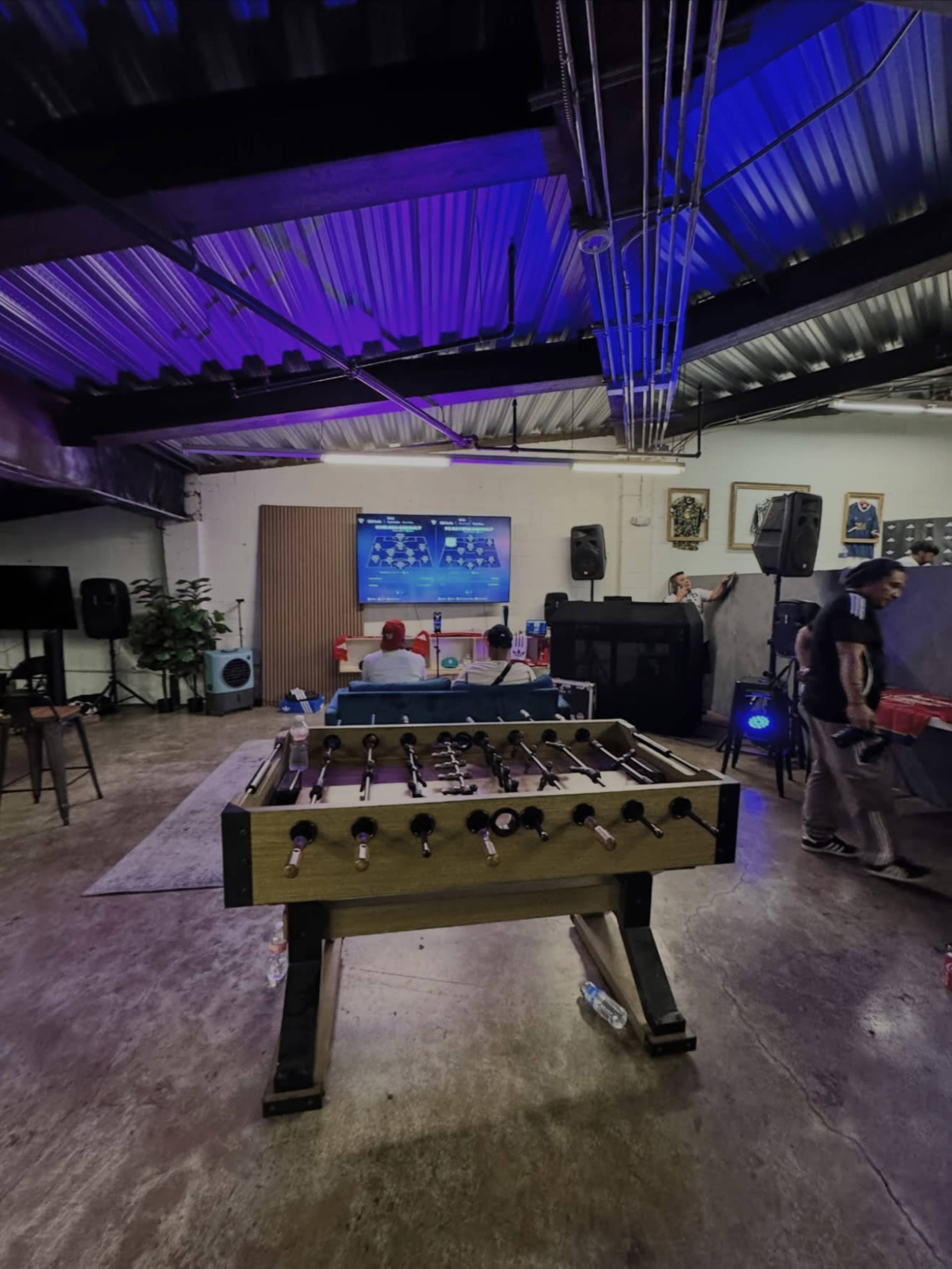 A foosball table is positioned in the foreground, with two people seated at a table in the background, watching a screen in a dimly lit room adorned with speakers and artwork.