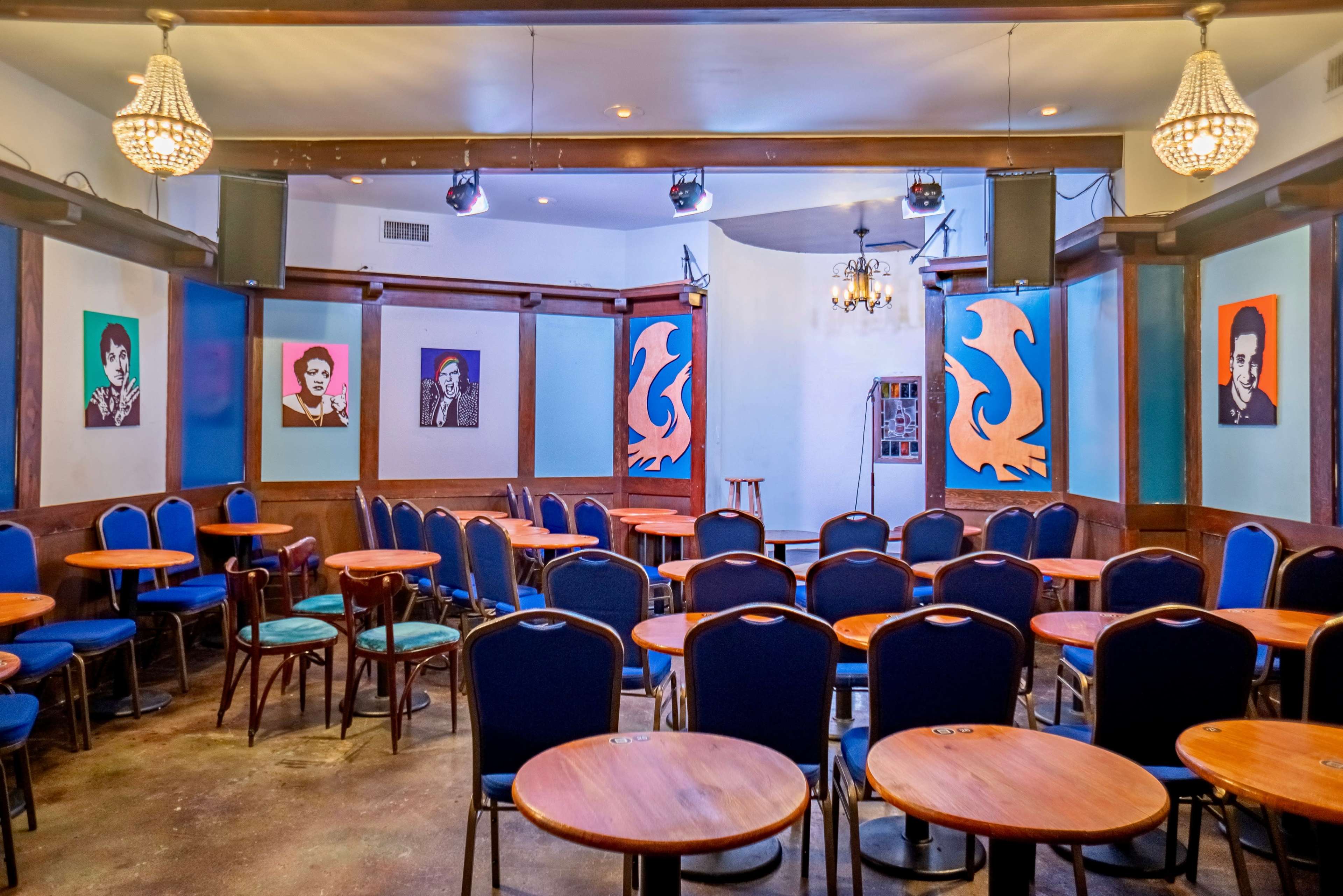 A small performance venue with round wooden tables and blue chairs, featuring colorful artwork on the walls.