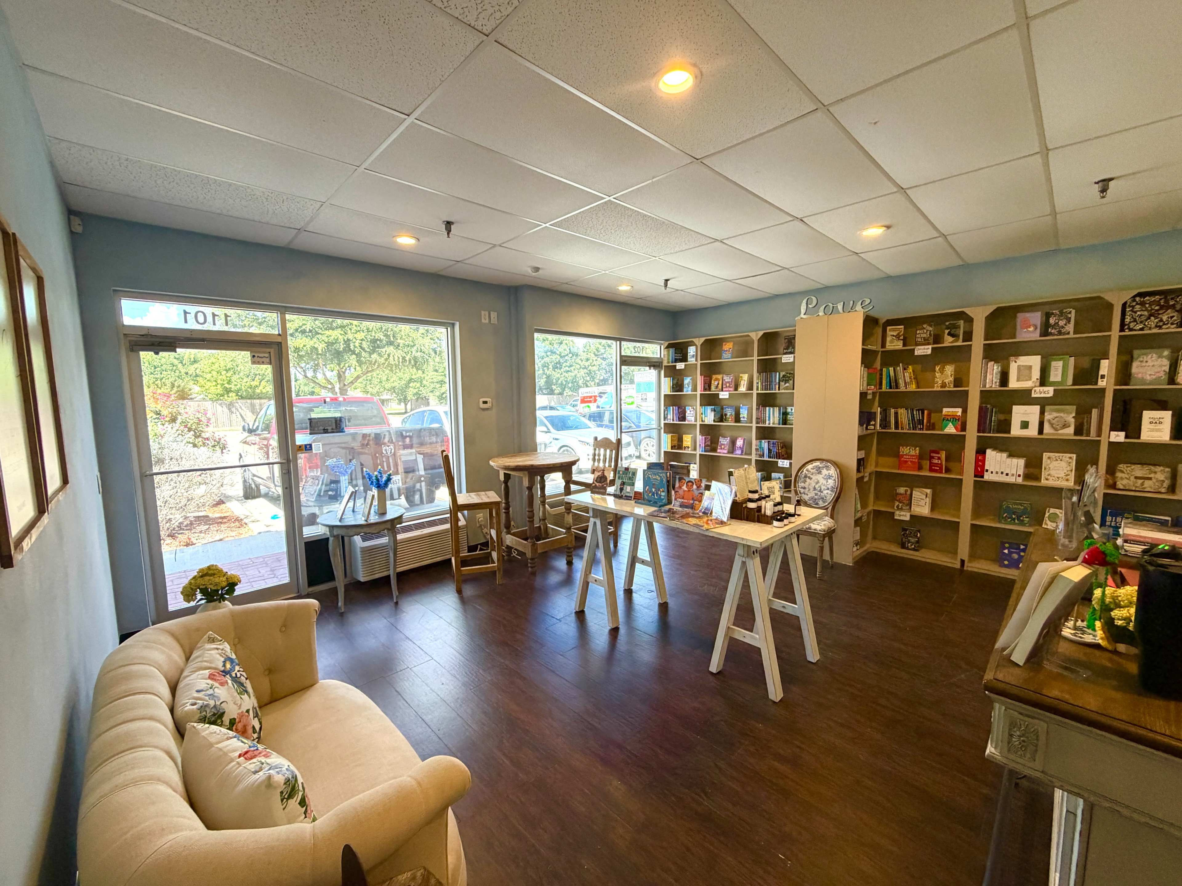 The image shows a cozy shop interior with a light blue wall, wooden flooring, a sofa, tables, and shelves filled with various products.
