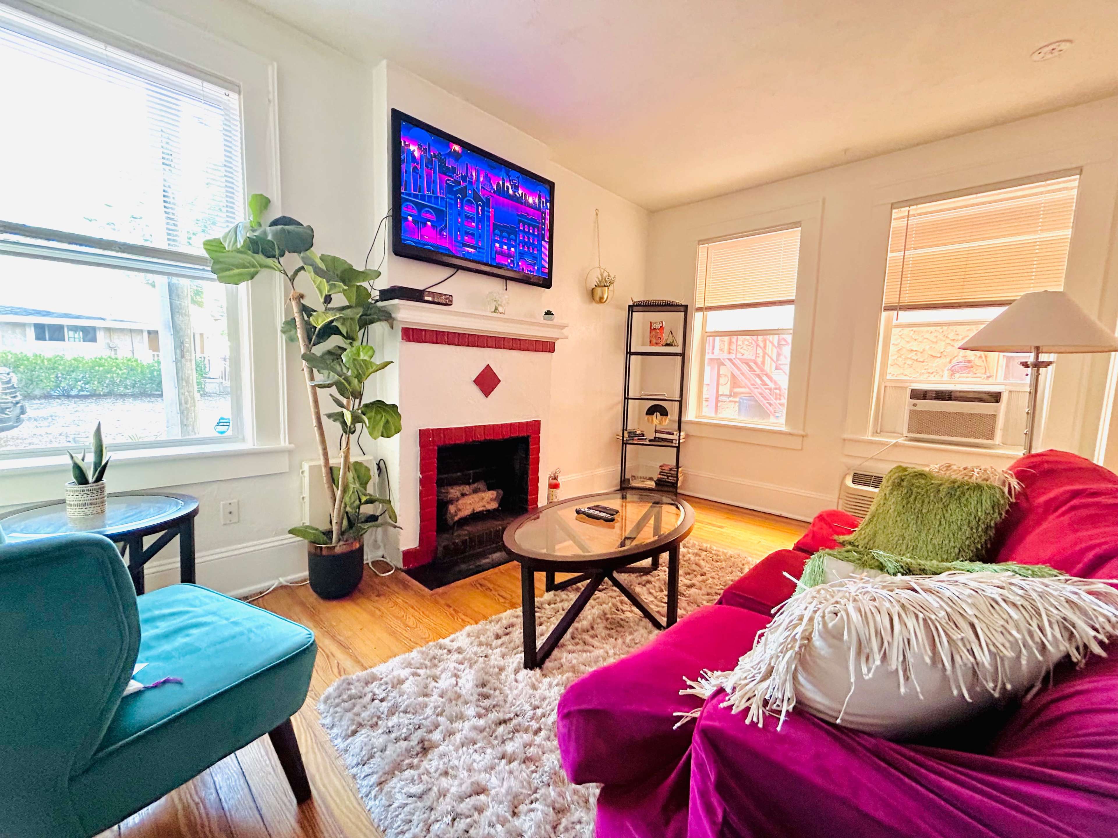 The living room features a green chair, a red couch with pillows, a round coffee table, and a fireplace with a mounted TV above it.