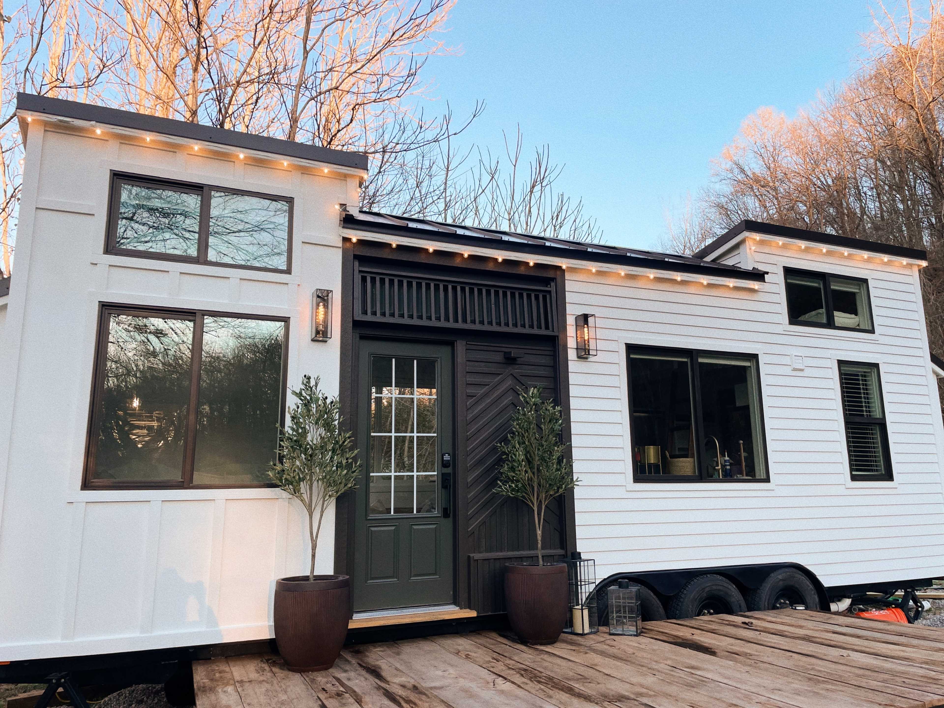 A modern tiny house on wheels, featuring a combination of white and dark siding, large windows, and a green front door, set against a backdrop of trees.