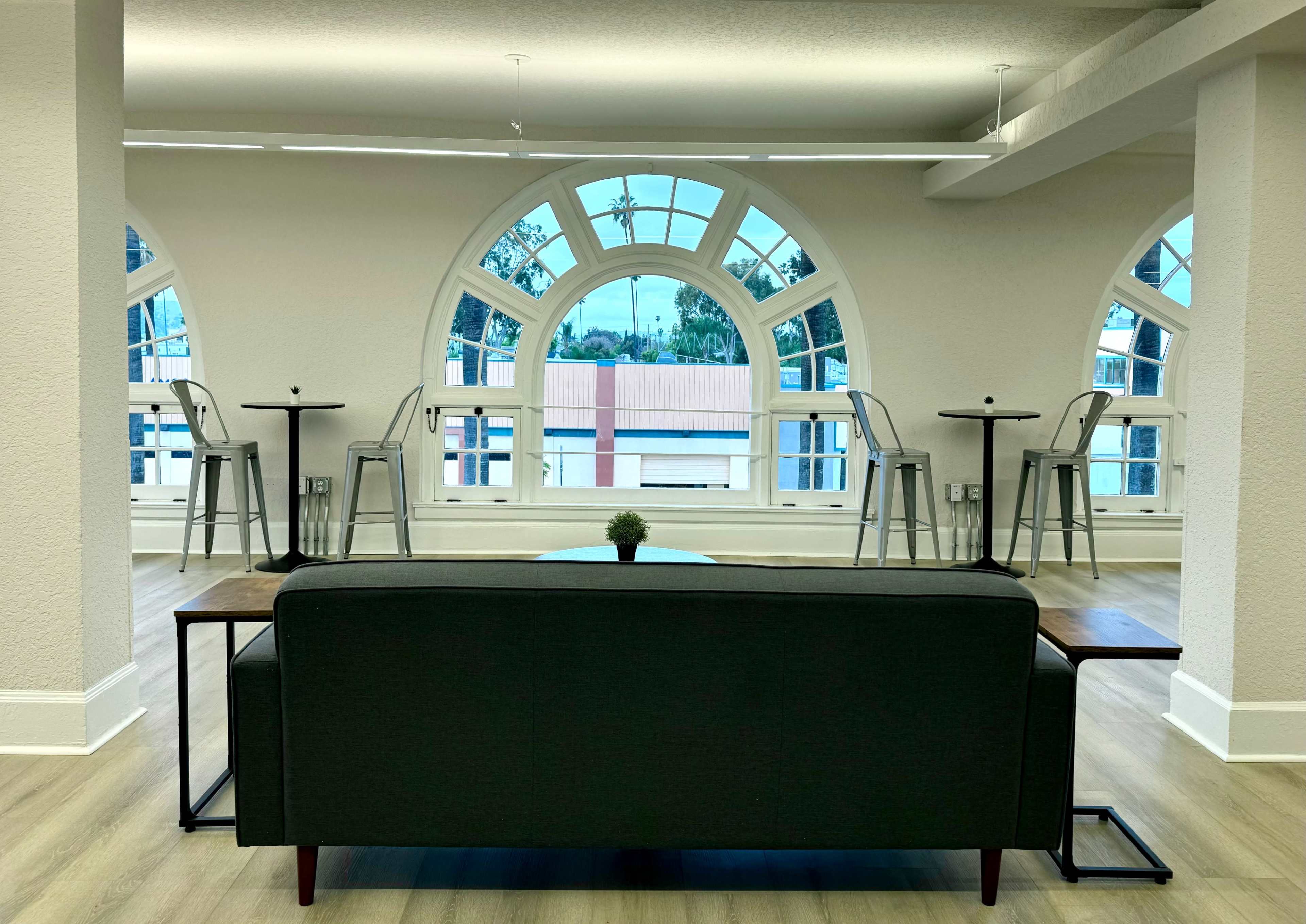 A modern seating area features a dark couch facing large arched windows, with high tables and stools arranged nearby.