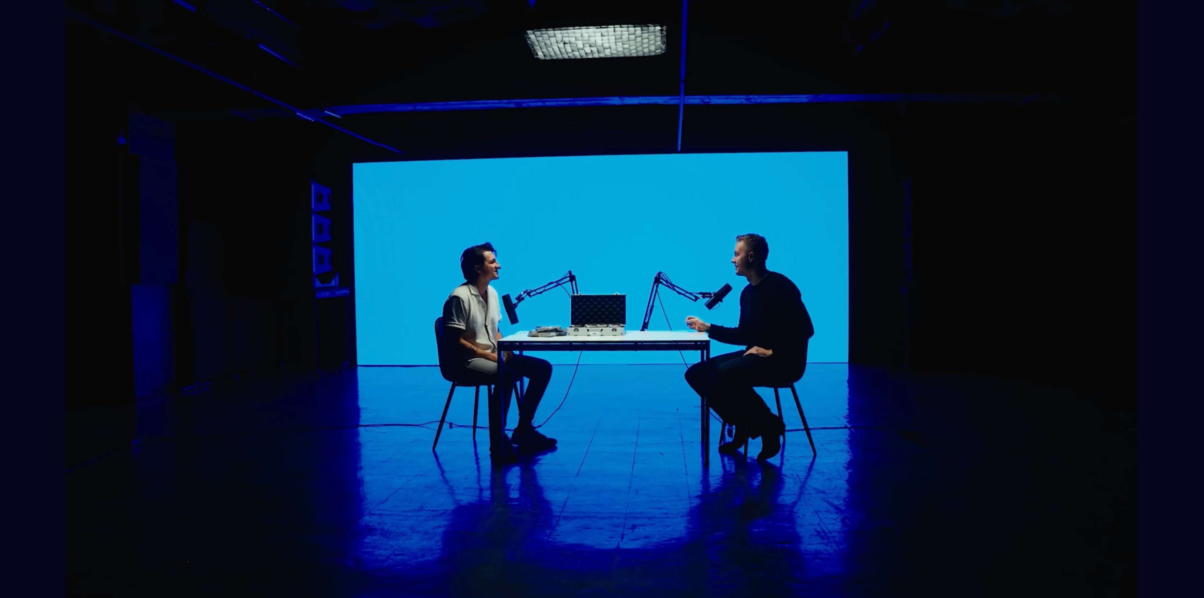 Two individuals sit across from each other at a table with microphones, facing a large blue screen in a dimly lit studio.
