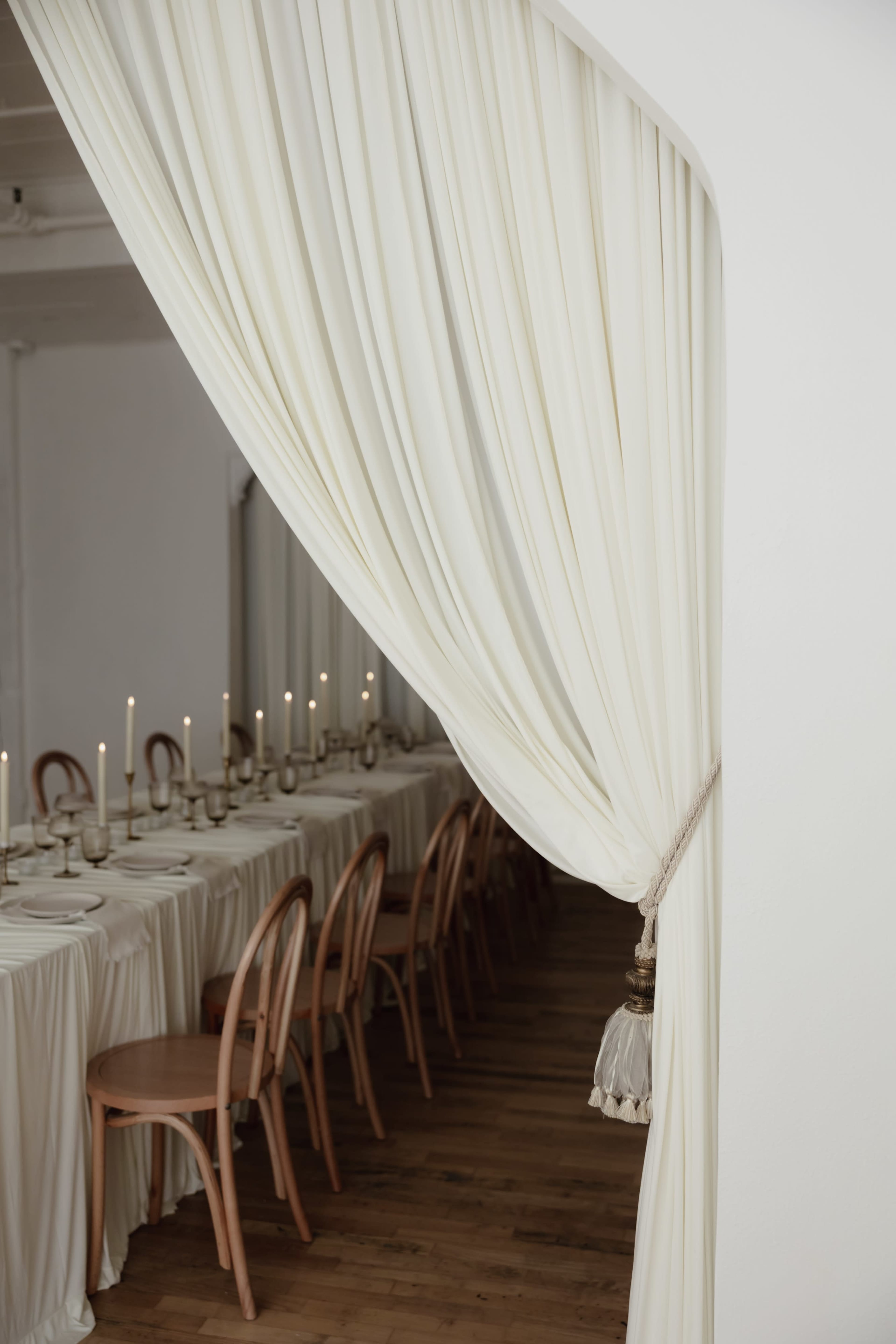 A long, elegantly set dining table with candles is partially revealed through a draped white curtain.