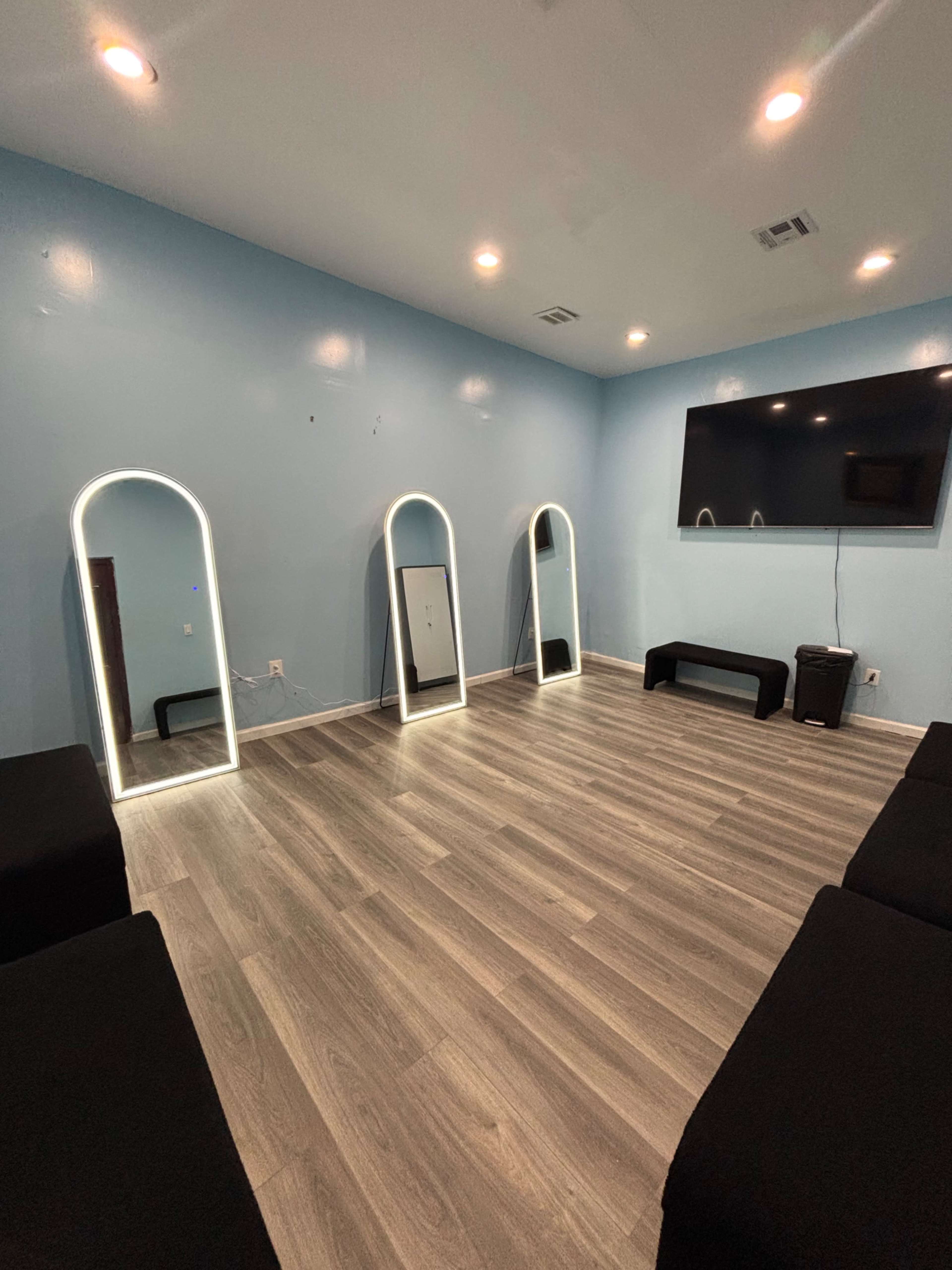 The room features three illuminated mirrors against a light blue wall, a large television mounted on the wall, and dark seating arrangements on a wooden floor.