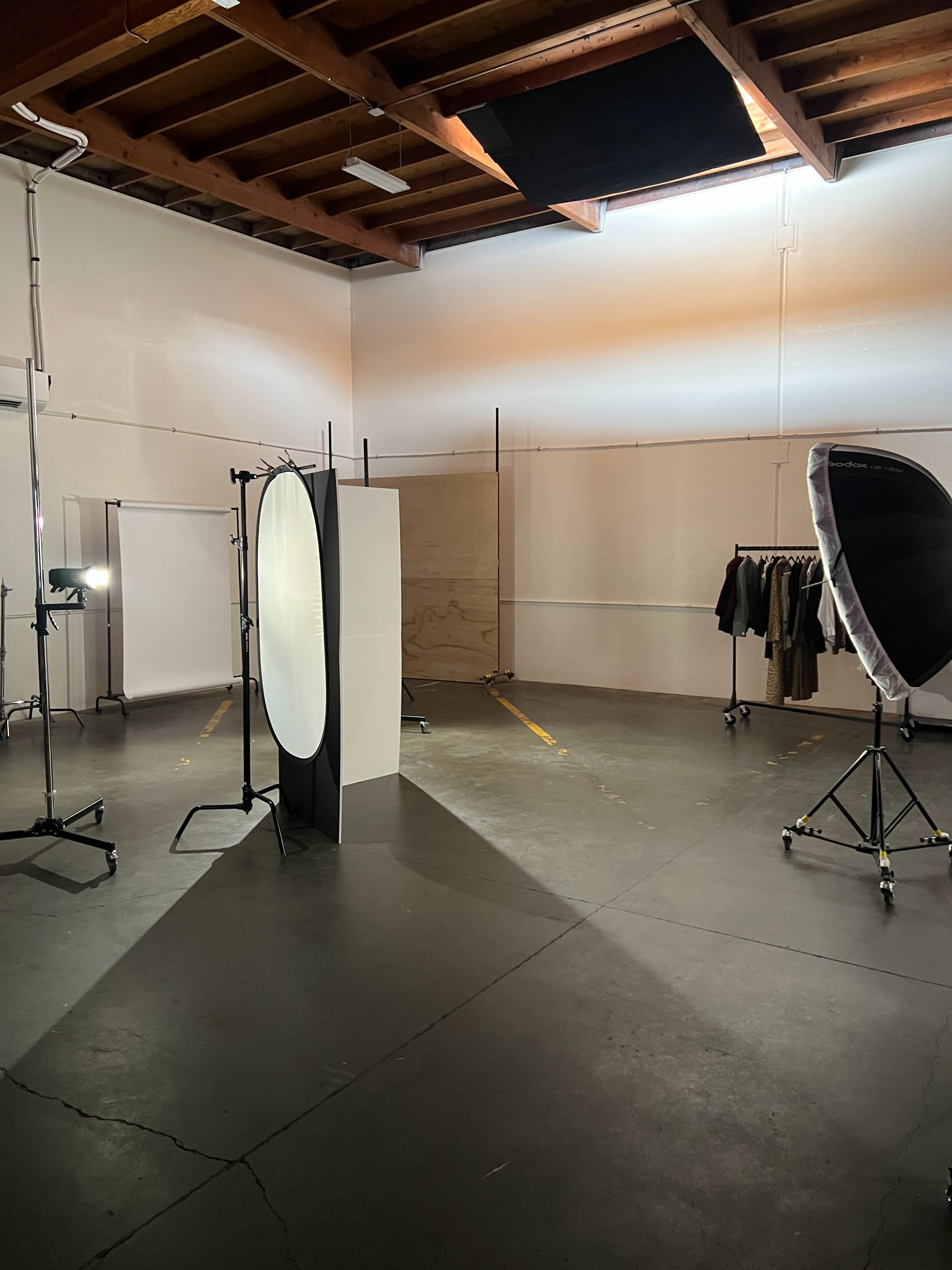 The image shows a spacious photography studio with various lighting equipment, a backdrop, and clothing racks arranged around a blank stand.