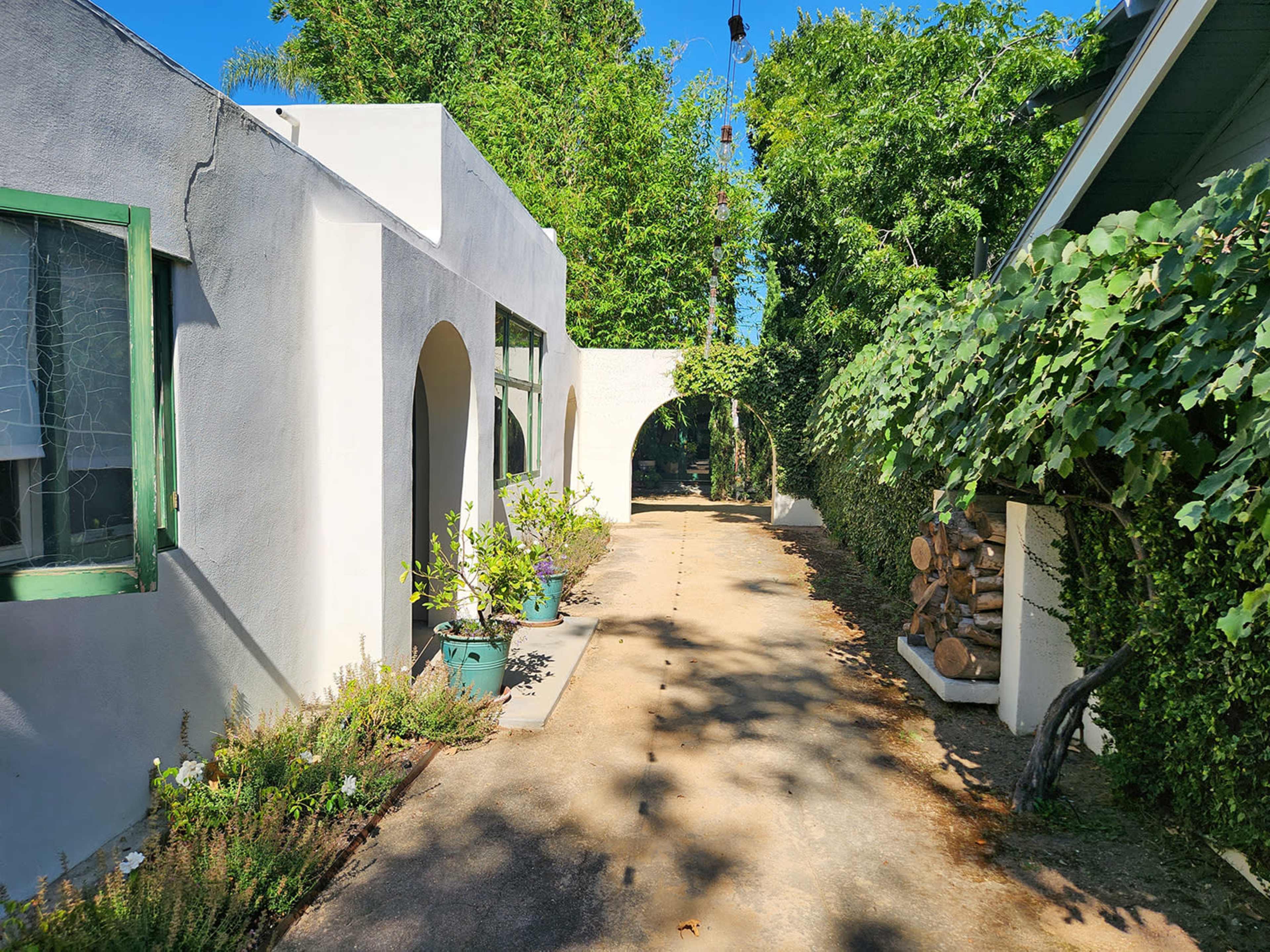 A narrow path leads between two white buildings, lined with potted plants and greenery on either side.