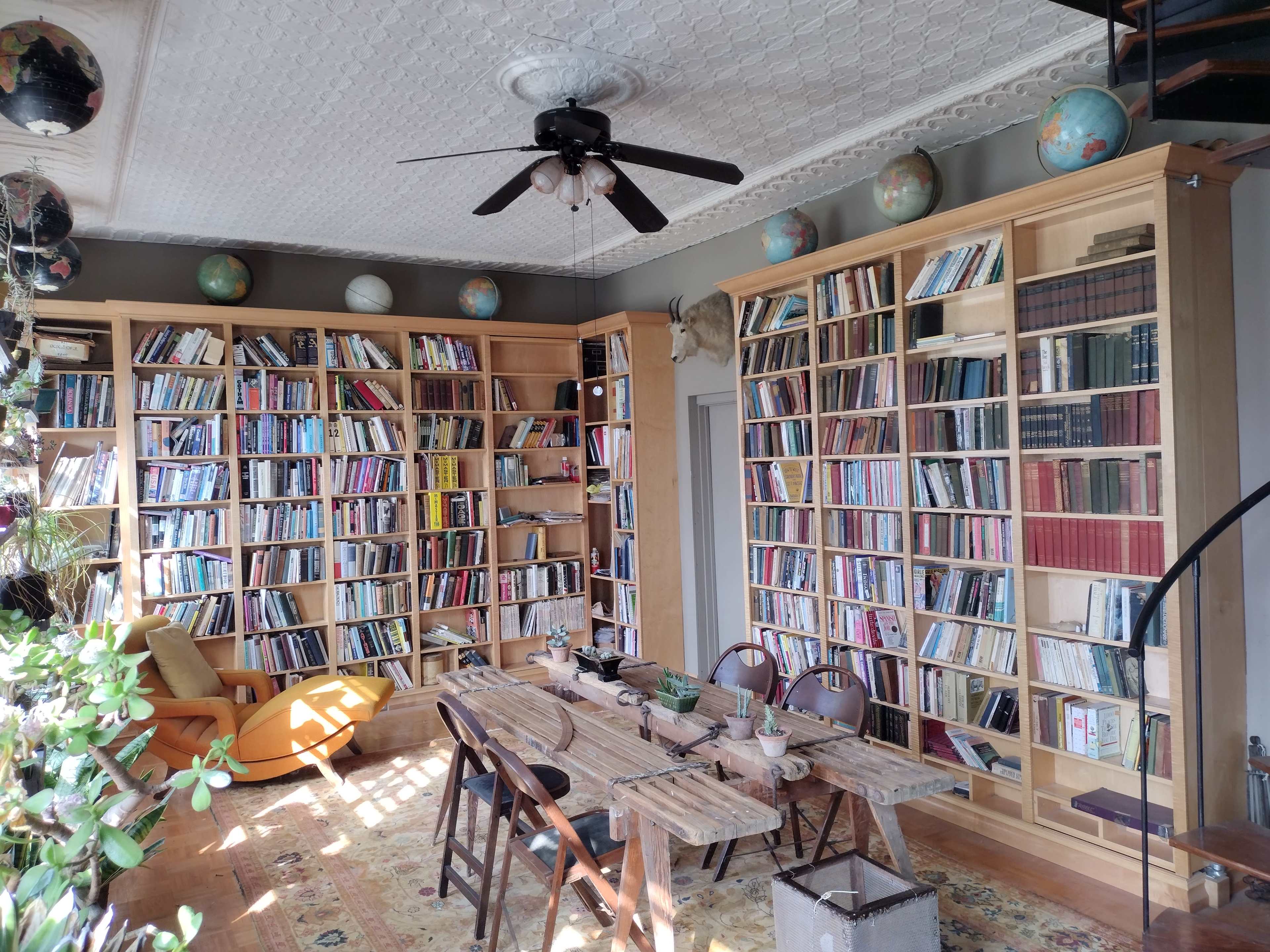 The image shows a cozy library with wooden bookshelves filled with books, a wooden table, and chairs, surrounded by plants and globes on the ceiling.