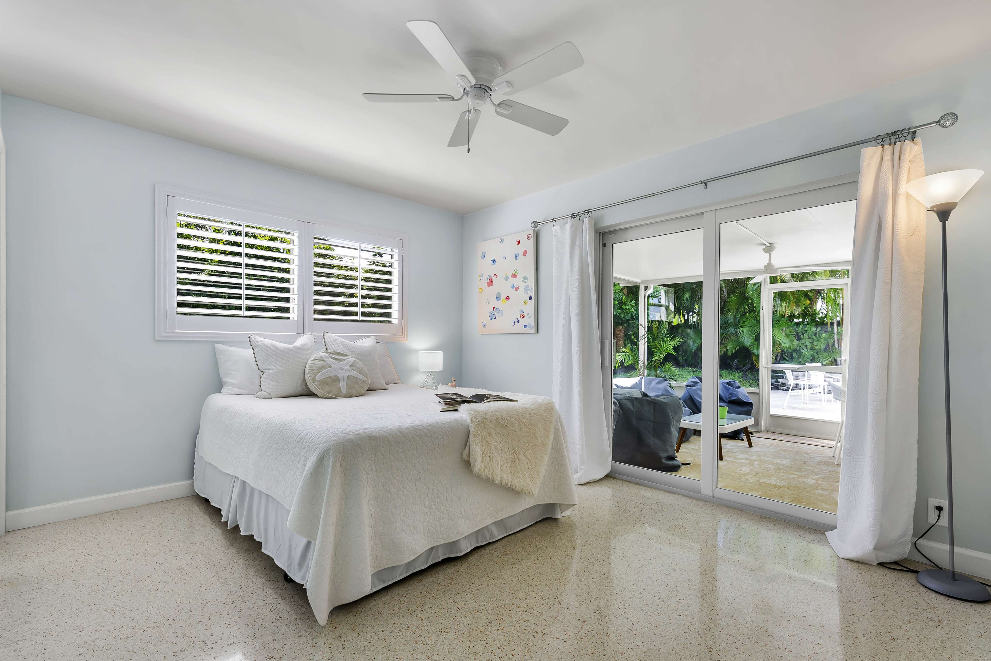 The image shows a light-colored bedroom featuring a white bed, large windows with shutters, and a sliding glass door leading to a patio area.