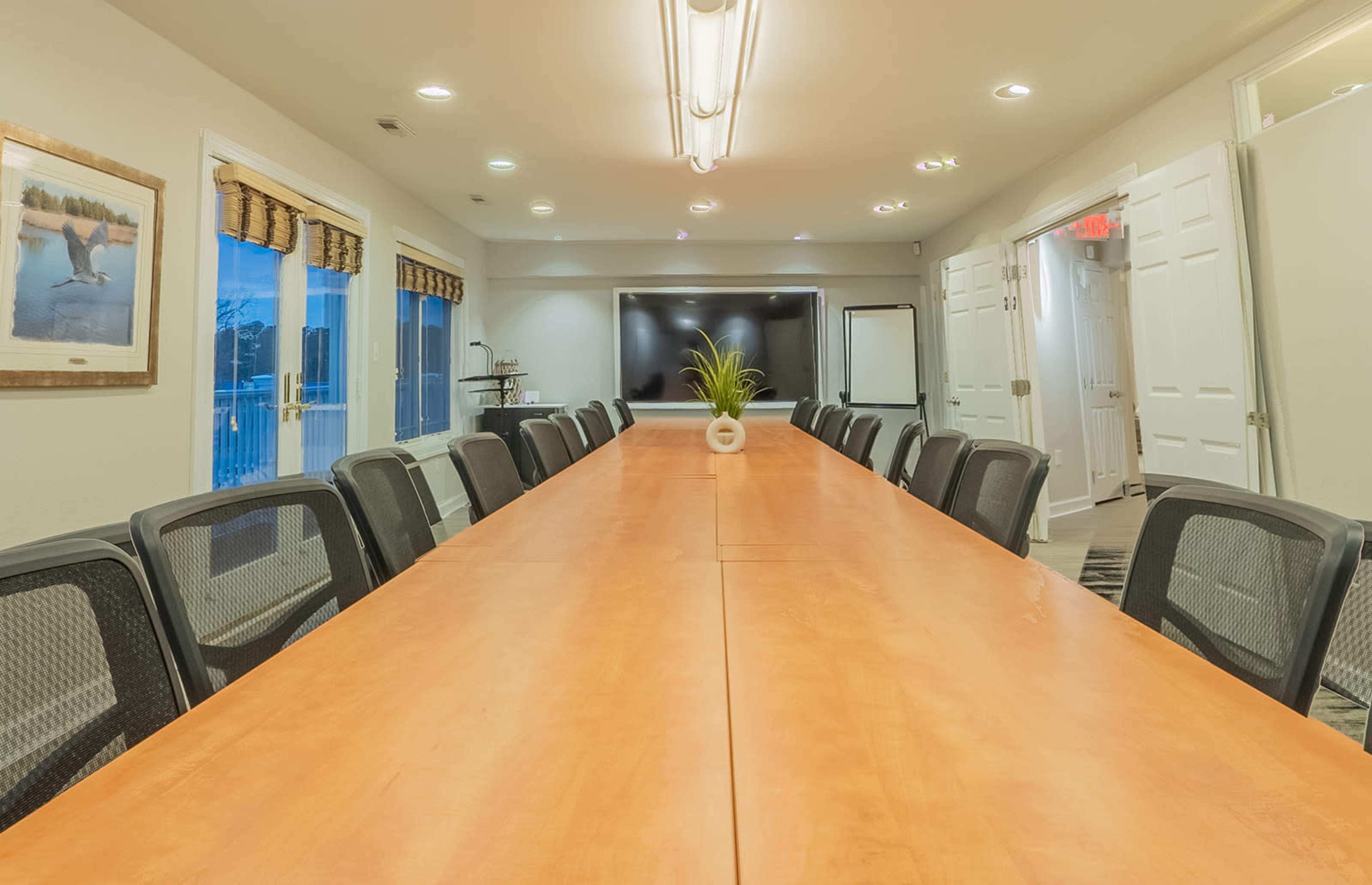 A large conference room with a wooden table, surrounded by office chairs, and equipped with a television and windows.