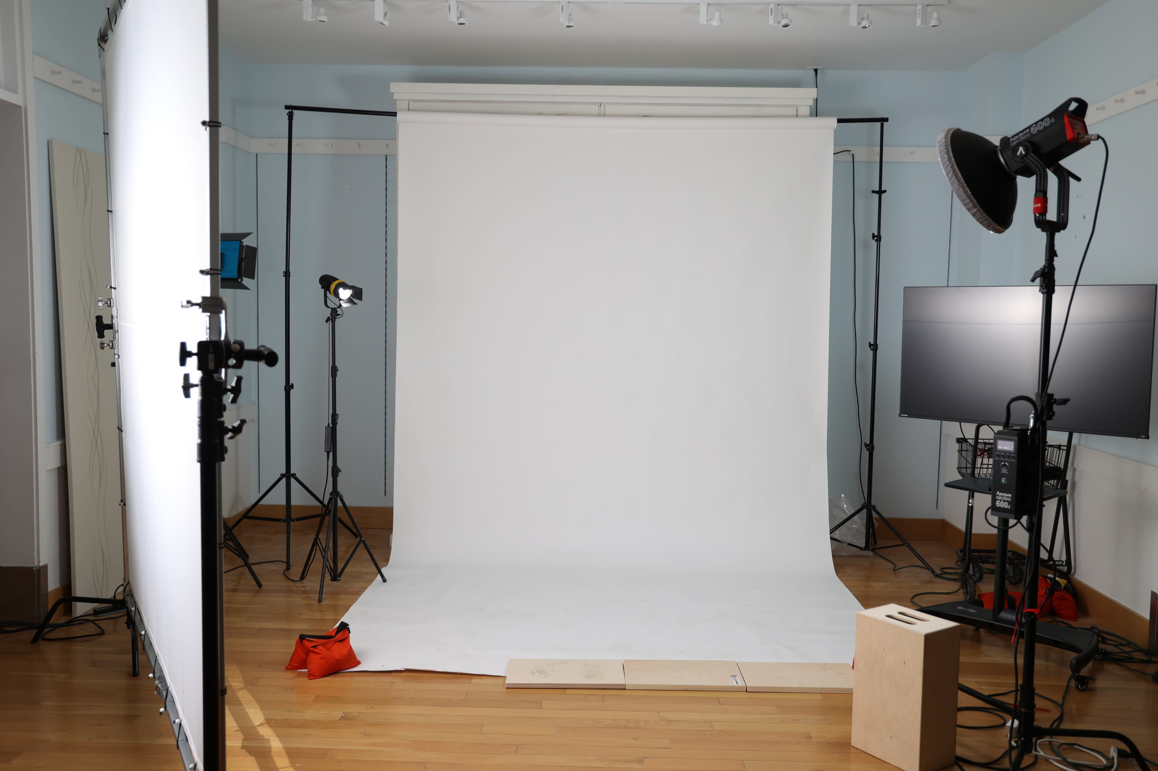 The image depicts a photography studio with a blank white backdrop, various lighting equipment, and a wooden platform on the floor.