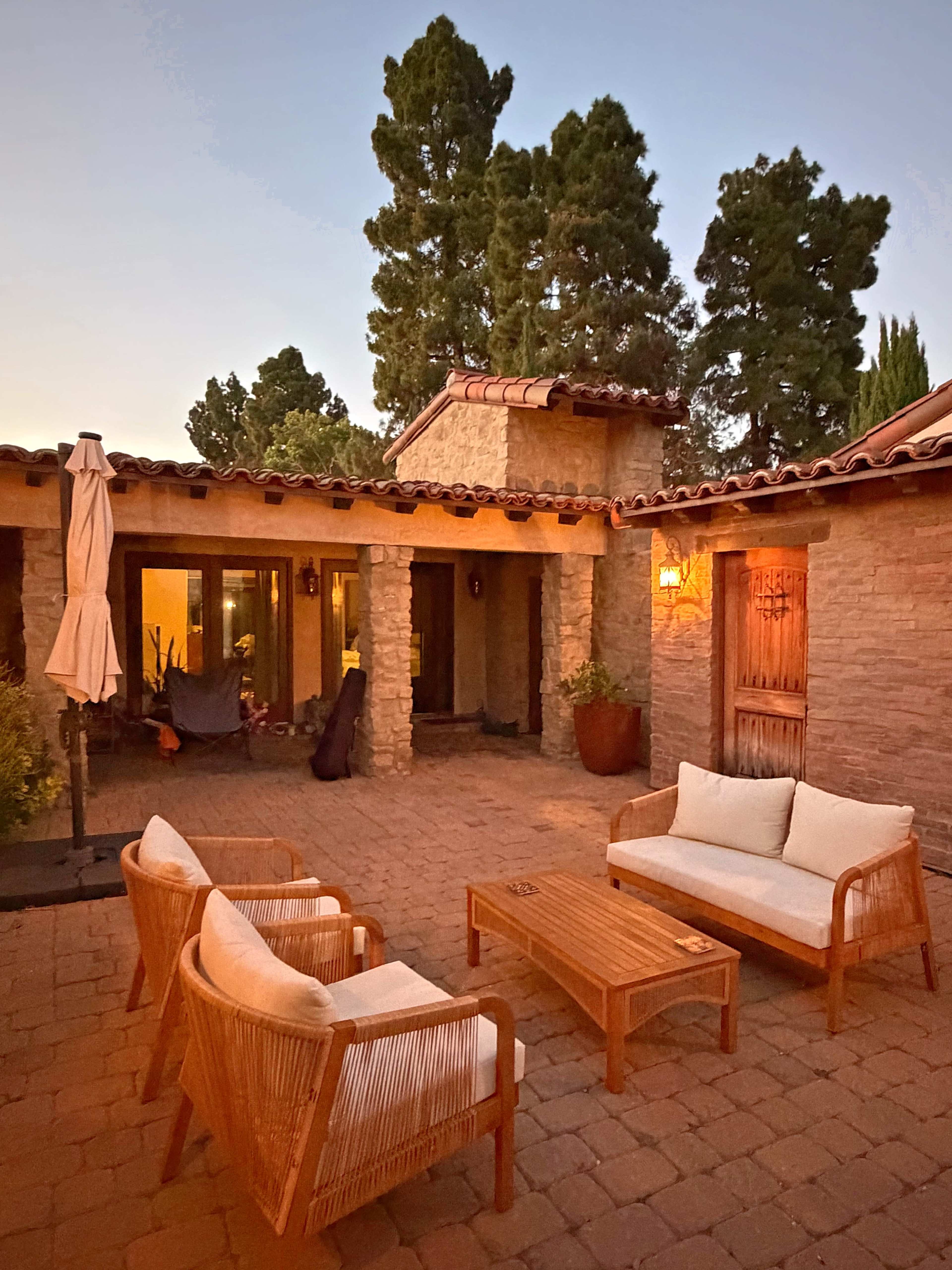 A cozy outdoor space features three wicker chairs and a wooden coffee table surrounded by a stone and stucco structure at dusk.