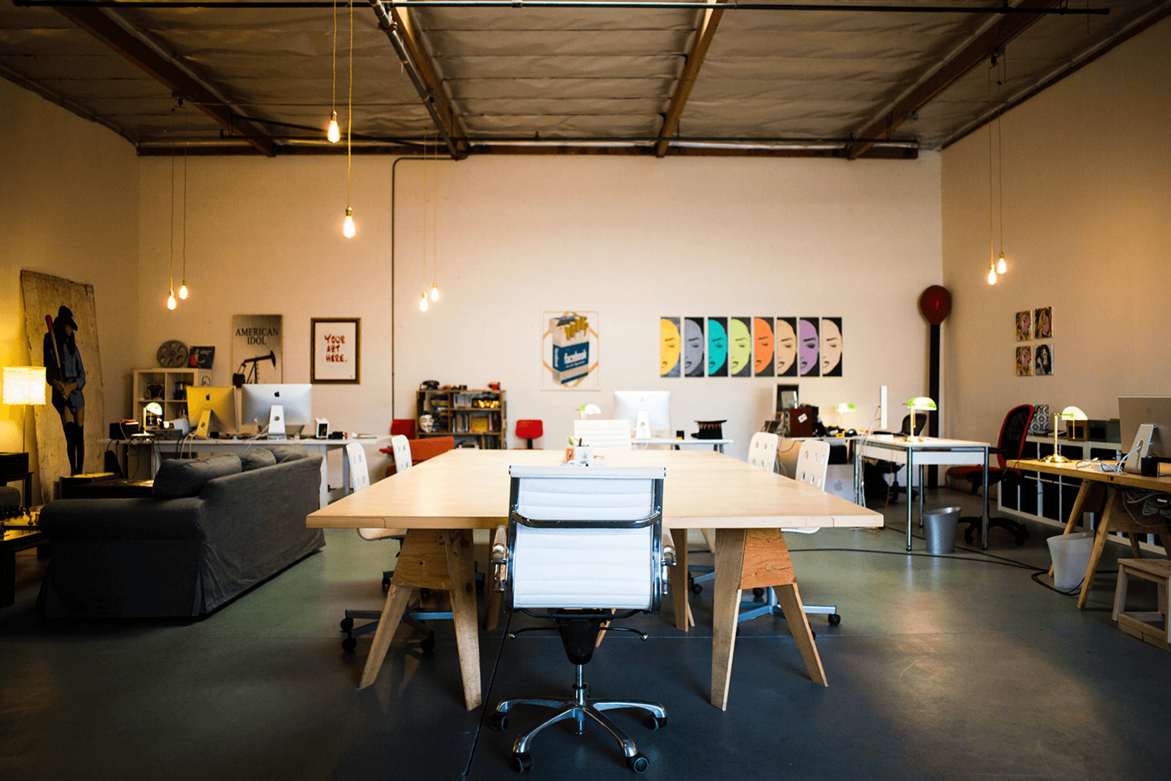 A large, open workspace features a central wooden table surrounded by chairs and various workstations equipped with computers, illuminated by hanging lights.
